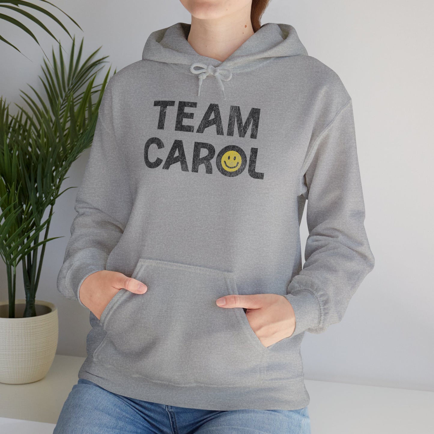 Woman wearing heather gray unisex Heavy Blend pullover hoodie sweatshirt with TEAM CAROL vintage distressed smiley graphic