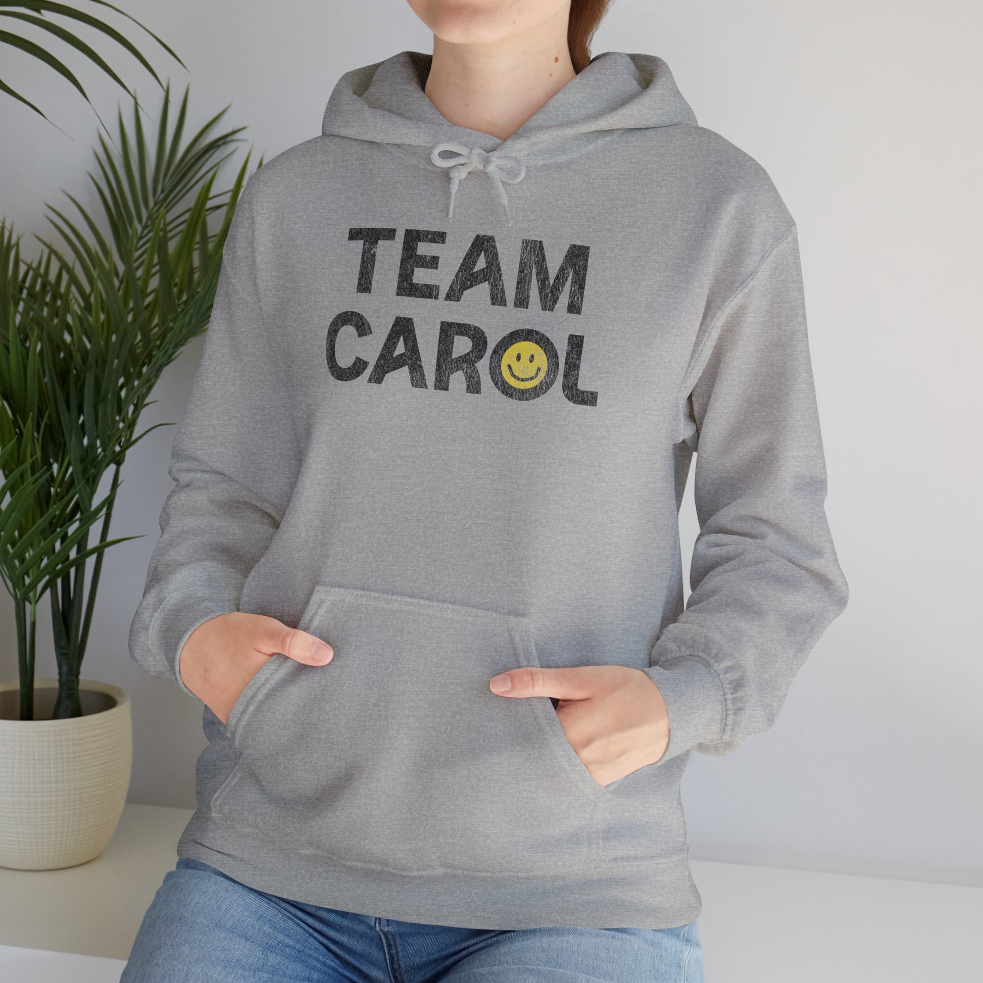 Woman wearing heather gray unisex Heavy Blend pullover hoodie sweatshirt with TEAM CAROL vintage distressed smiley graphic