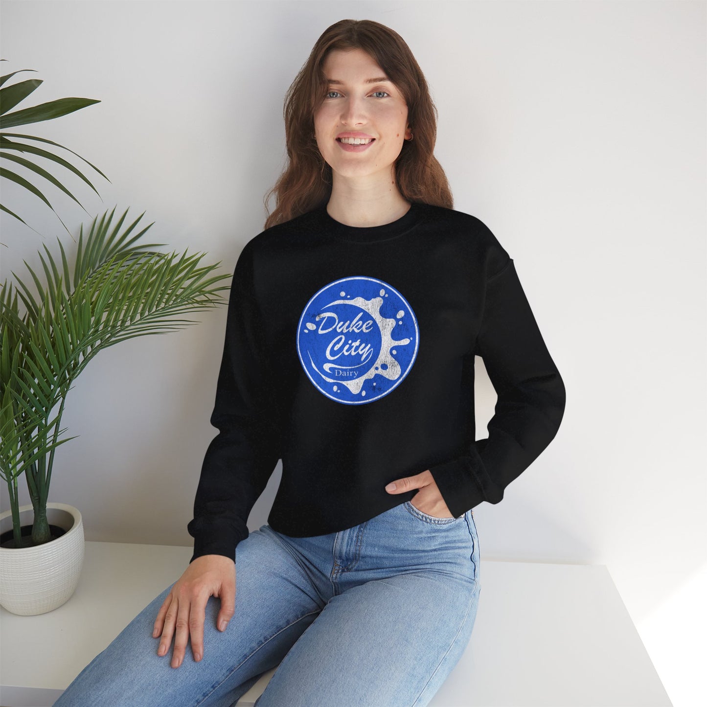 Woman wearing a black sweatshirt with 'Duke City' logo design from Pluribus, sitting on a white surface with a plant in the background.