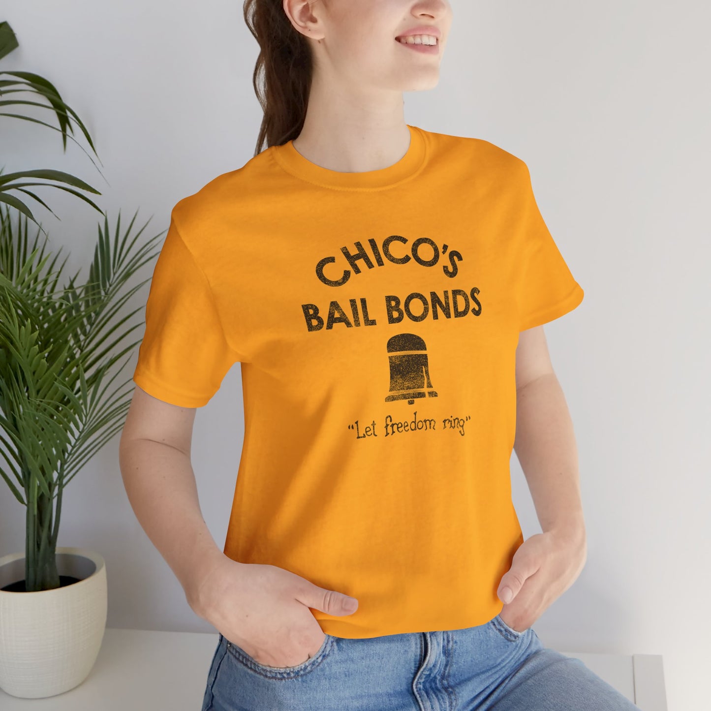 female wearing gold t-shirt with distressed Chico’s Bail Bonds logo and “Let freedom ring” slogan from The Bad News Bears movie.