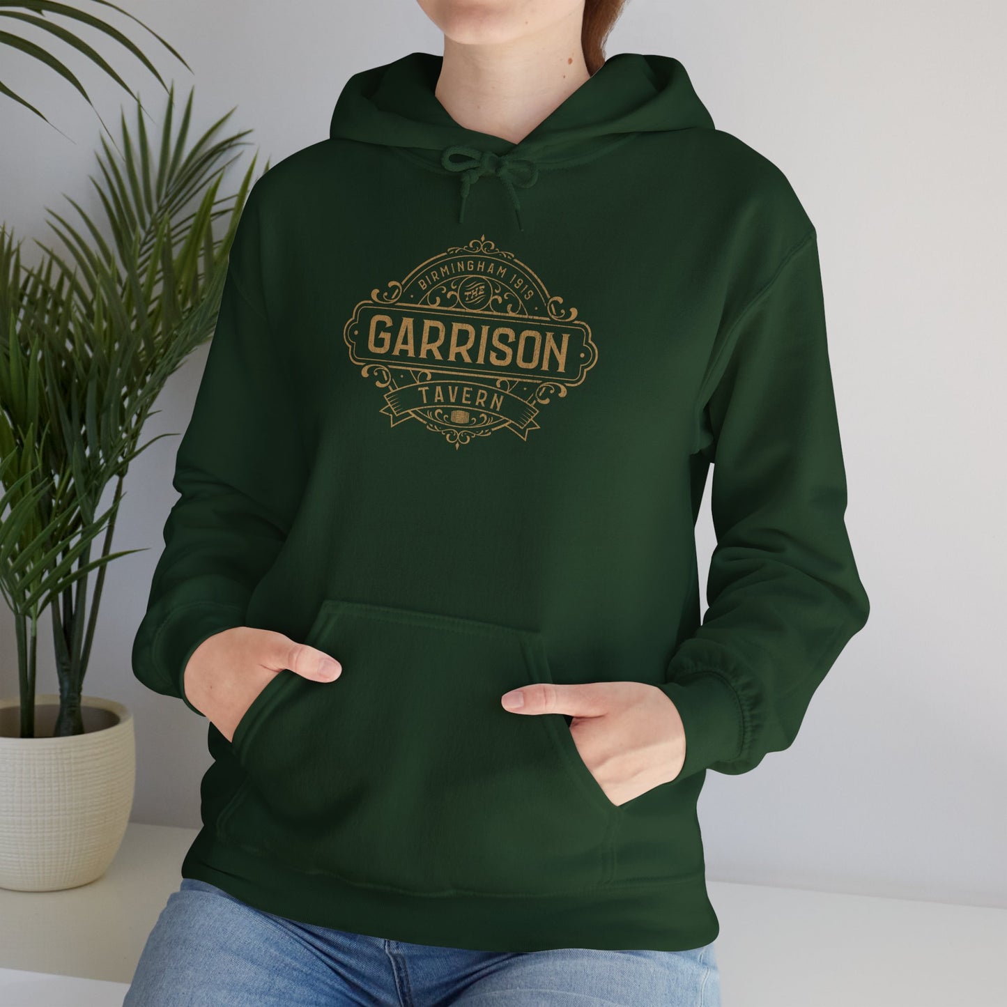 Female model wearing a forest green Soft Unisex Hoodie Heavy Blend Hooded Sweatshirts featuring the Garrison Tavern Bar Logo Graphic inspired by the TV show and movie Peaky Blinders in a vintage distressed style sold by ScriptedBrand.