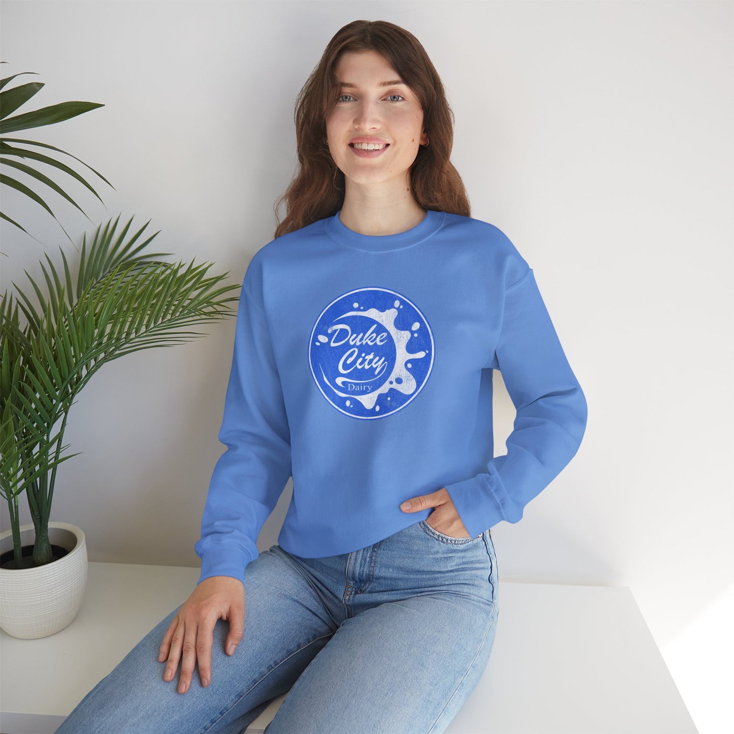 Woman wearing a light blue sweatshirt with 'Duke City' logo design from Pluribus, sitting on a white surface with a plant in the background.