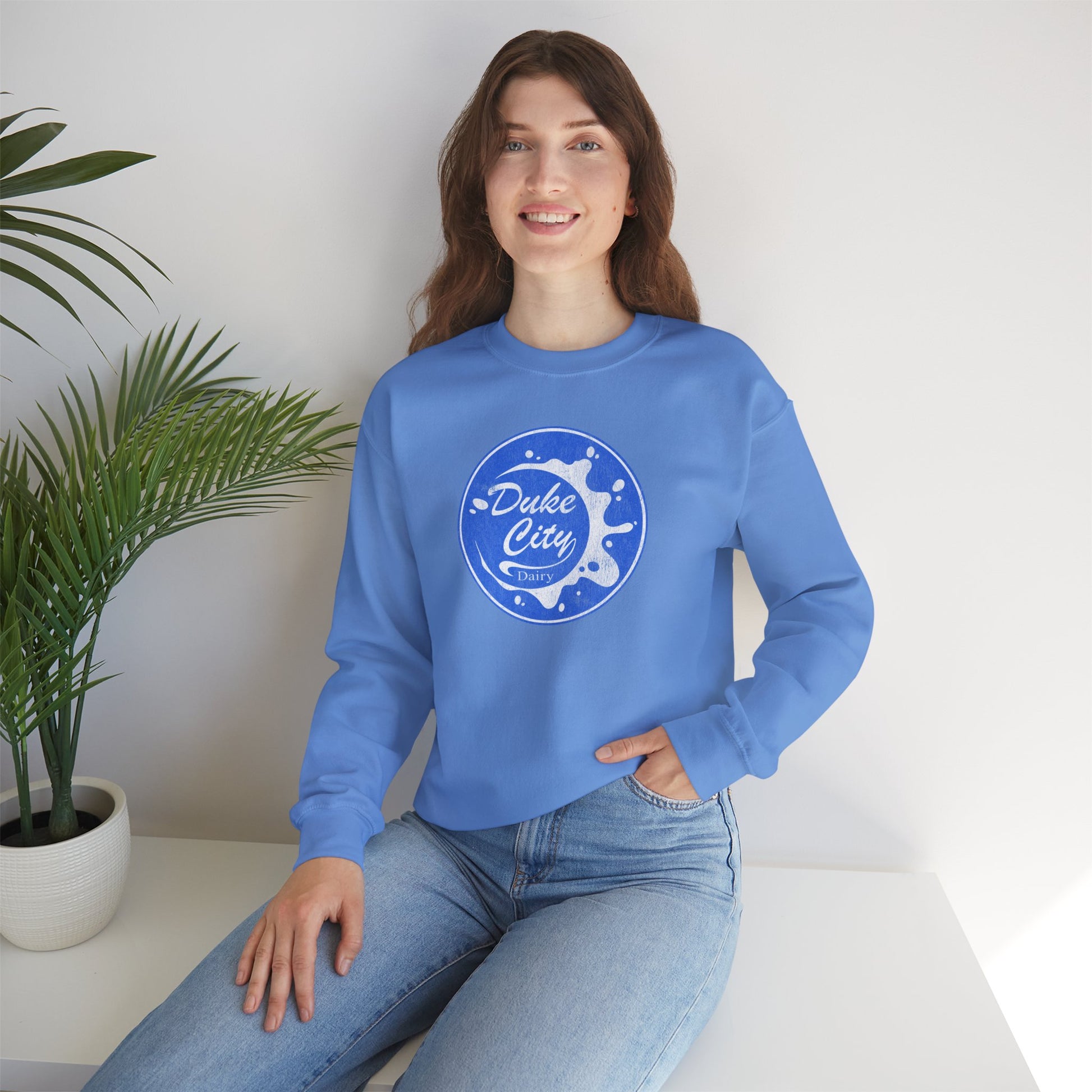 Woman wearing a light blue sweatshirt with 'Duke City' logo design from Pluribus, sitting on a white surface with a plant in the background.