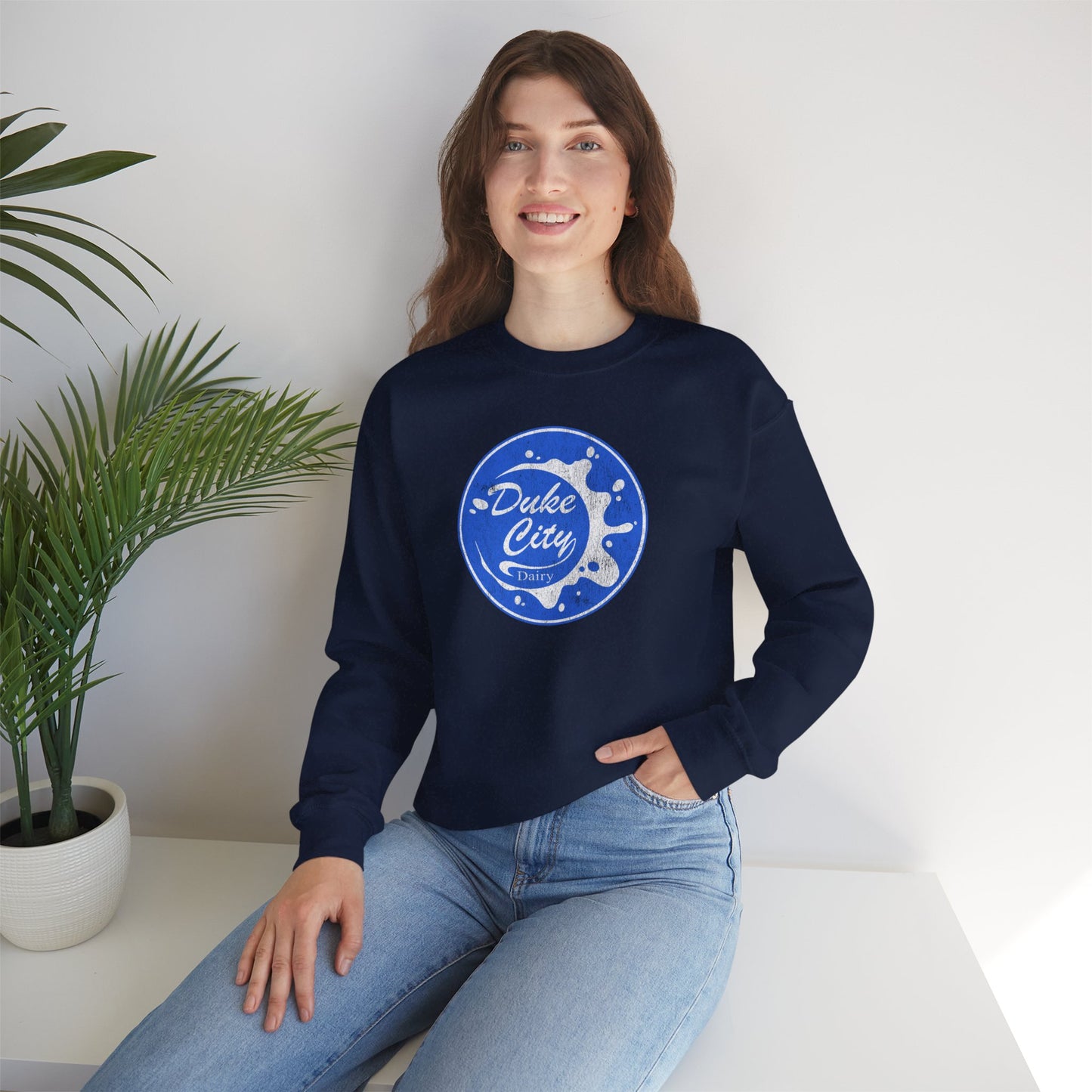 Woman wearing a navy blue sweatshirt with 'Duke City' logo design from Pluribus, sitting on a white surface with a plant in the background.