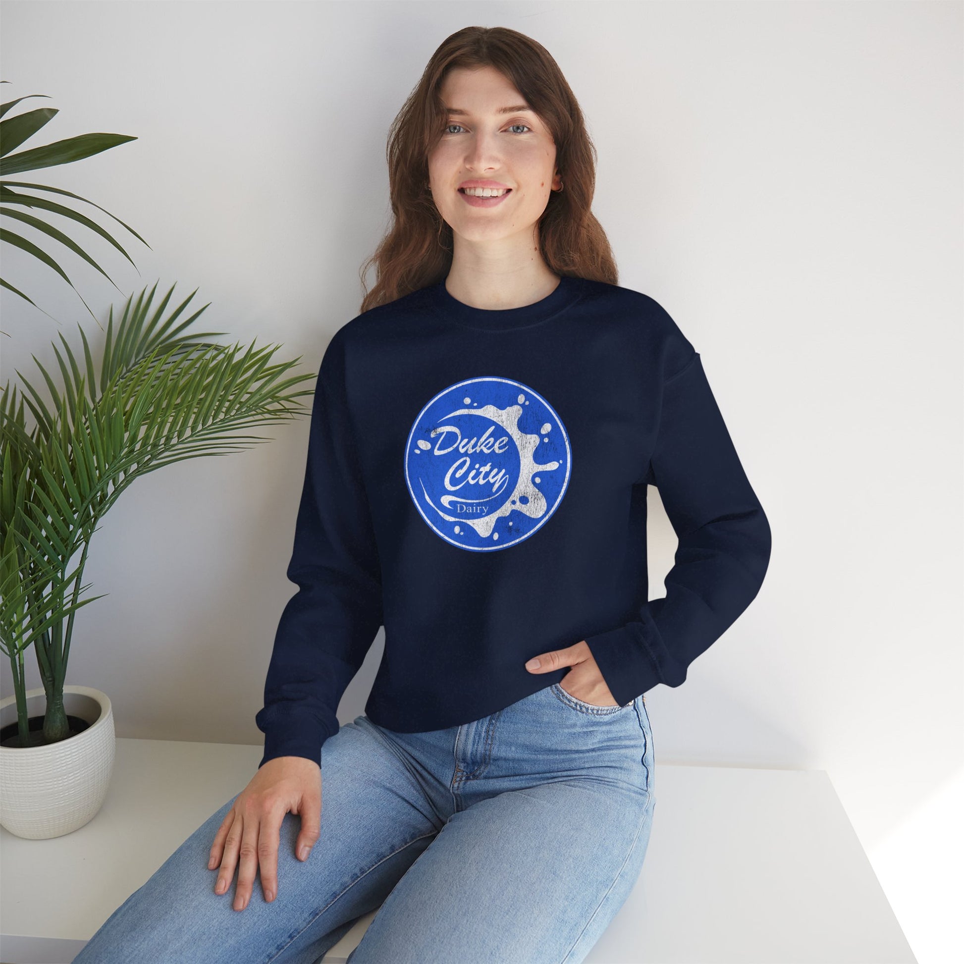 Woman wearing a navy blue sweatshirt with 'Duke City' logo design from Pluribus, sitting on a white surface with a plant in the background.
