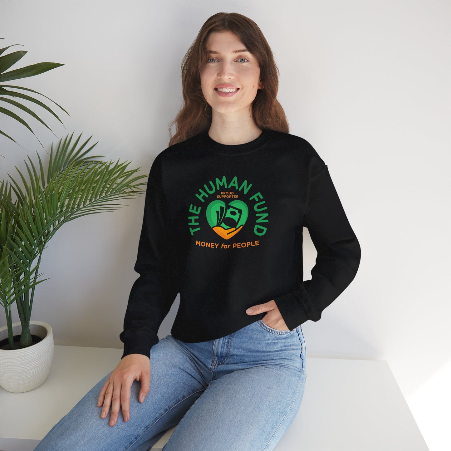 Person wearing a black sweatshirt with 'The Human Fund' logo sitting on a white surface with a plant in the background.