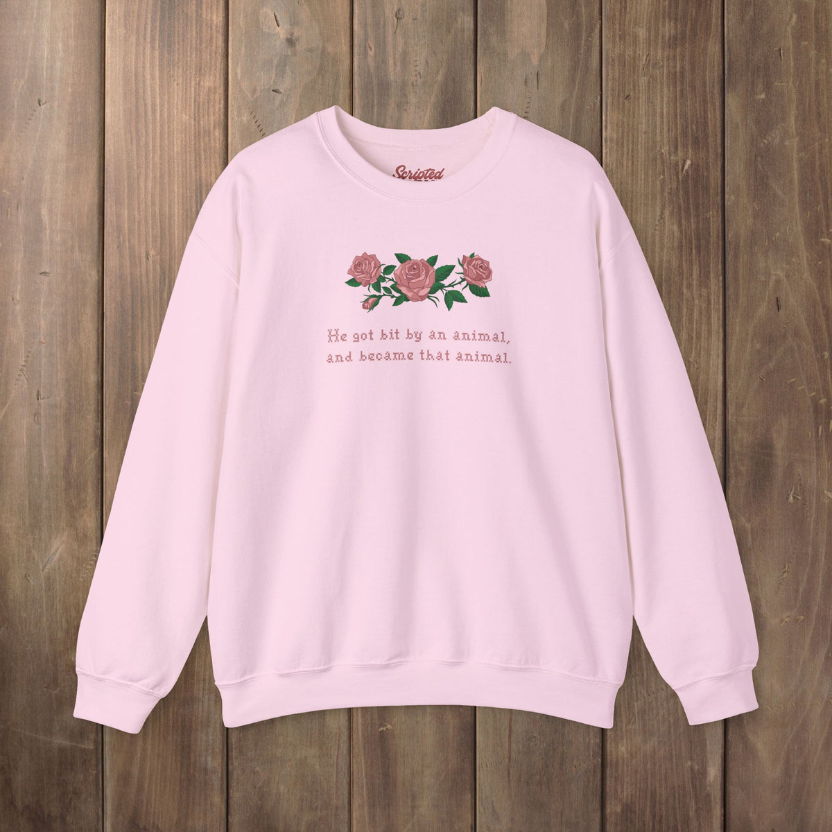 A light pink crewneck sweatshirt with a graphic of pink roses and green leaves on the chest, accompanied by the text 'He hit by an animal, and became that animal' in pink. The sweatshirt is laid flat on a rustic wooden plank background.
