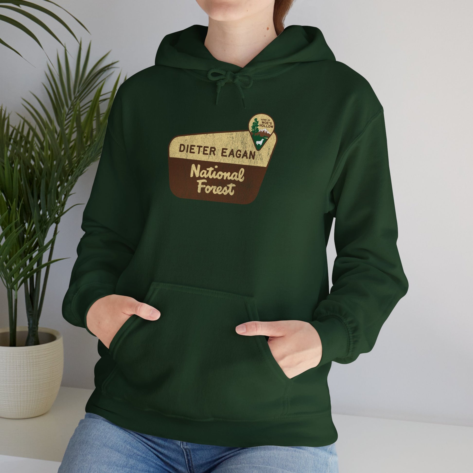 Female model wearing a Forest green colored Soft Unisex Hoodie Heavy Blend Hooded Sweatshirt featuring the logo graphic of the Dieter Eagan National Forest inspired by the TV show Severance in a vintage distressed ink-stamped worn-in style.