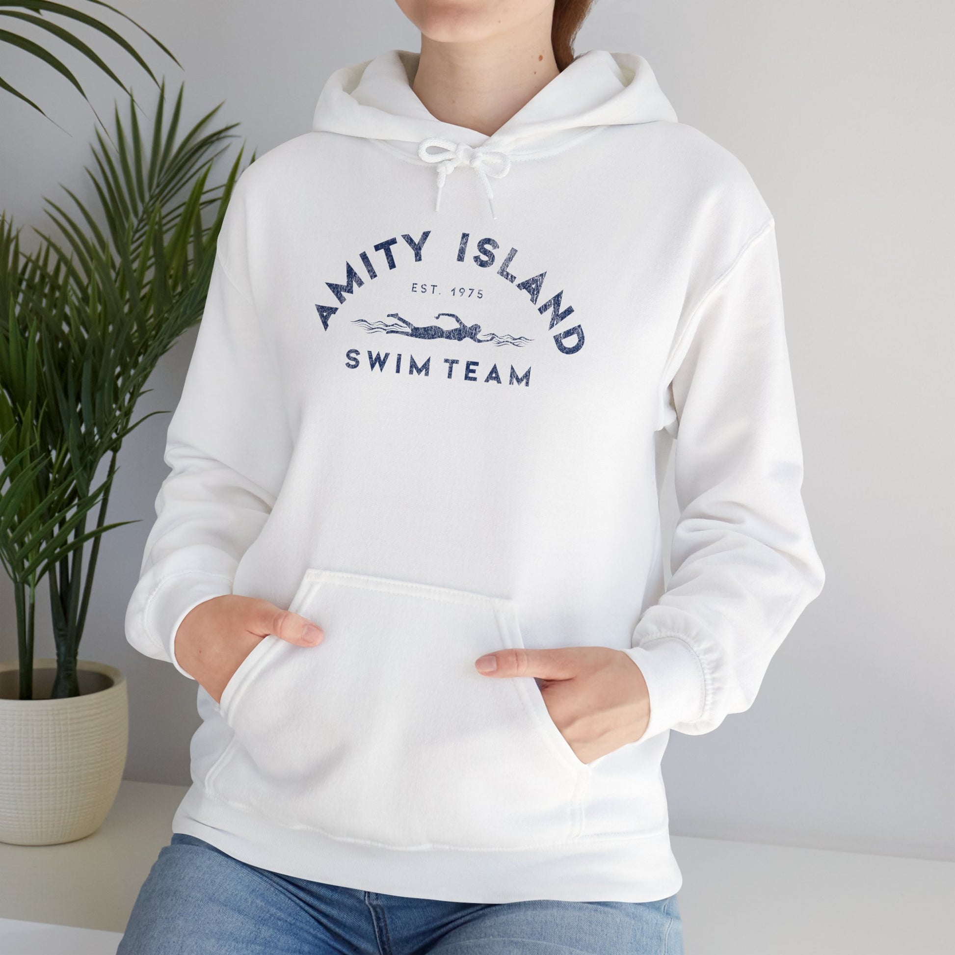 Female model wearing a white colored Soft Unisex Hoodie Heavy Blend Hooded Sweatshirt featuring an Amity Island Swim Team logo graphic, designed by ScriptedBrand, and  inspired by the movie Jaws in a vintage distressed style.