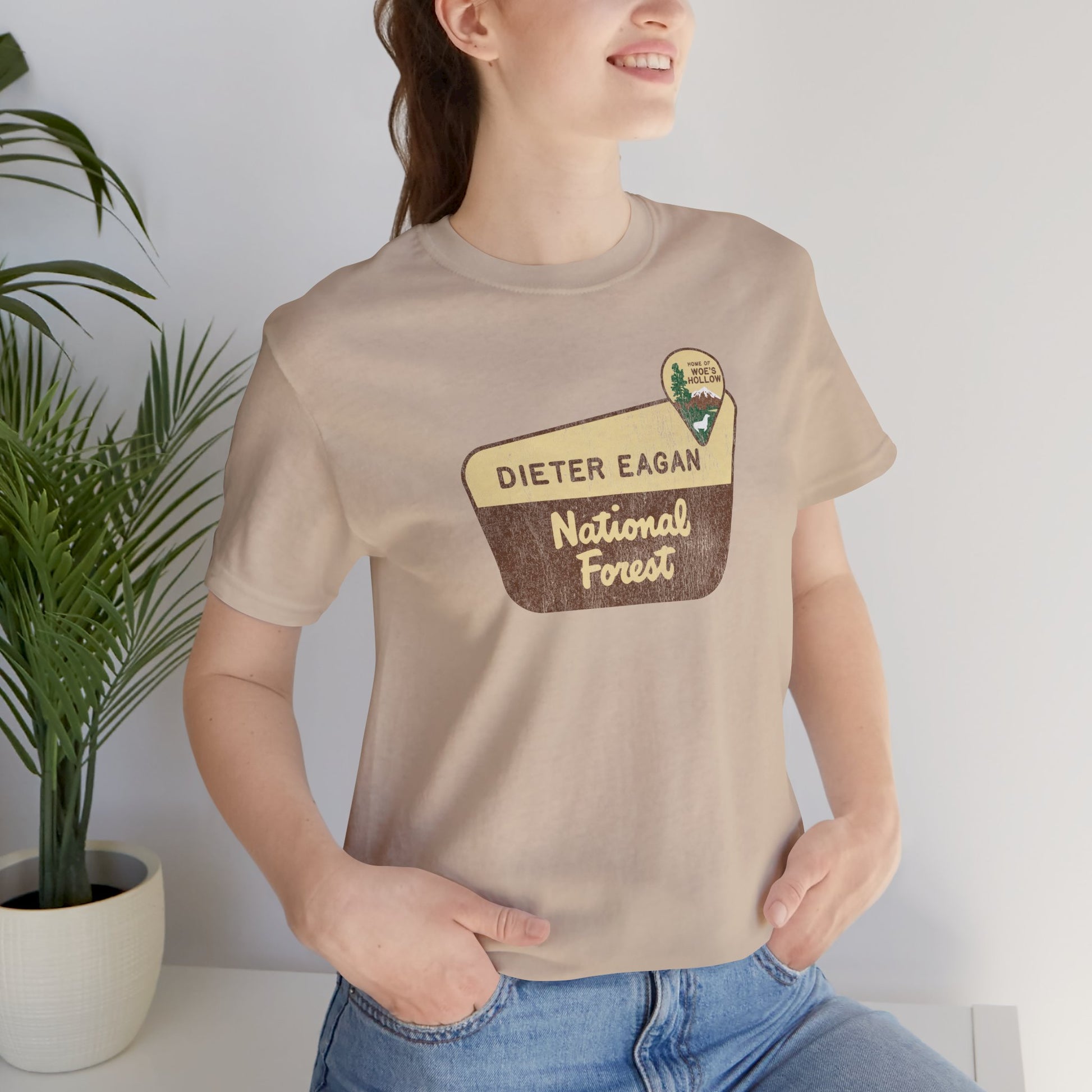 Female model wearing a tan colored soft Bella+Canvas short-sleeve t-shirt with the logo graphic of the Dieter Eagan National Forest inspired by the TV show Severance in a vintage distressed ink-stamped worn-in style.