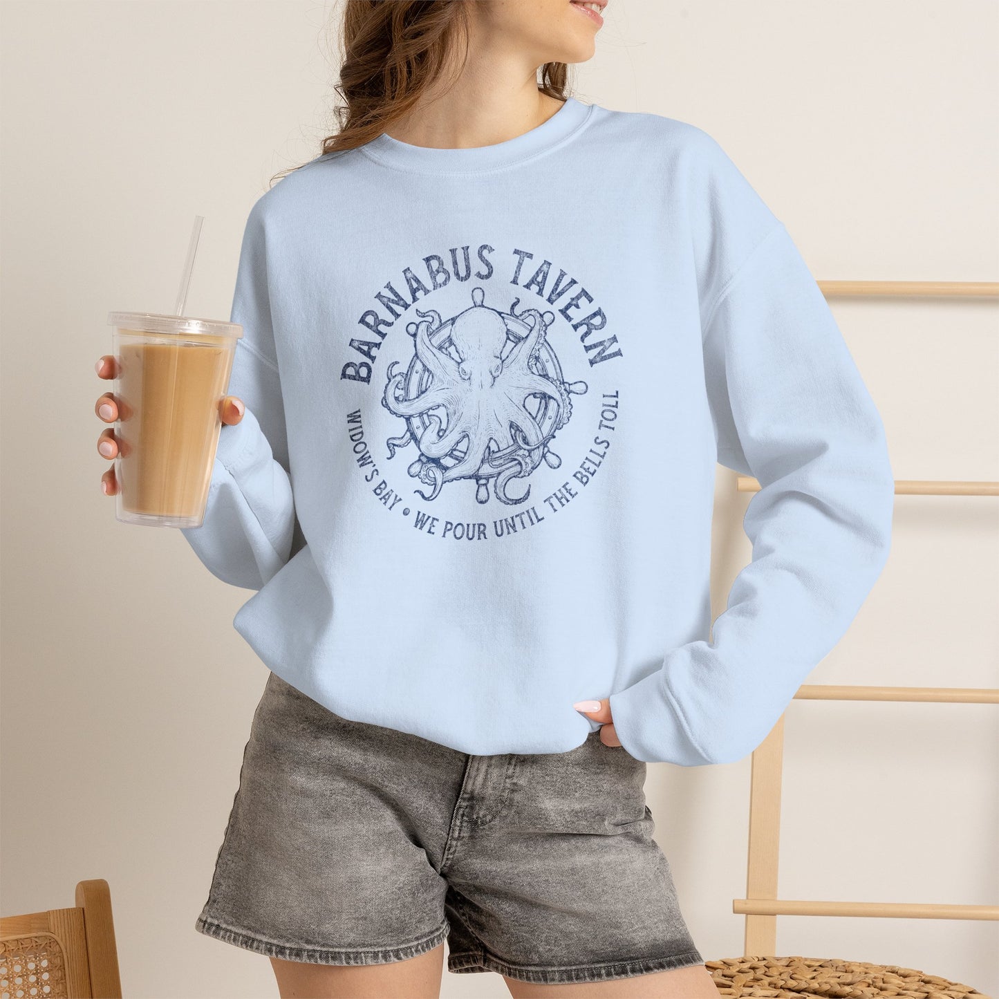 A woman wearing a light blue crewneck sweatshirt with a dark blue 'Barnabus Tavern' octopus graphic, paired with gray denim shorts and holding an iced coffee.