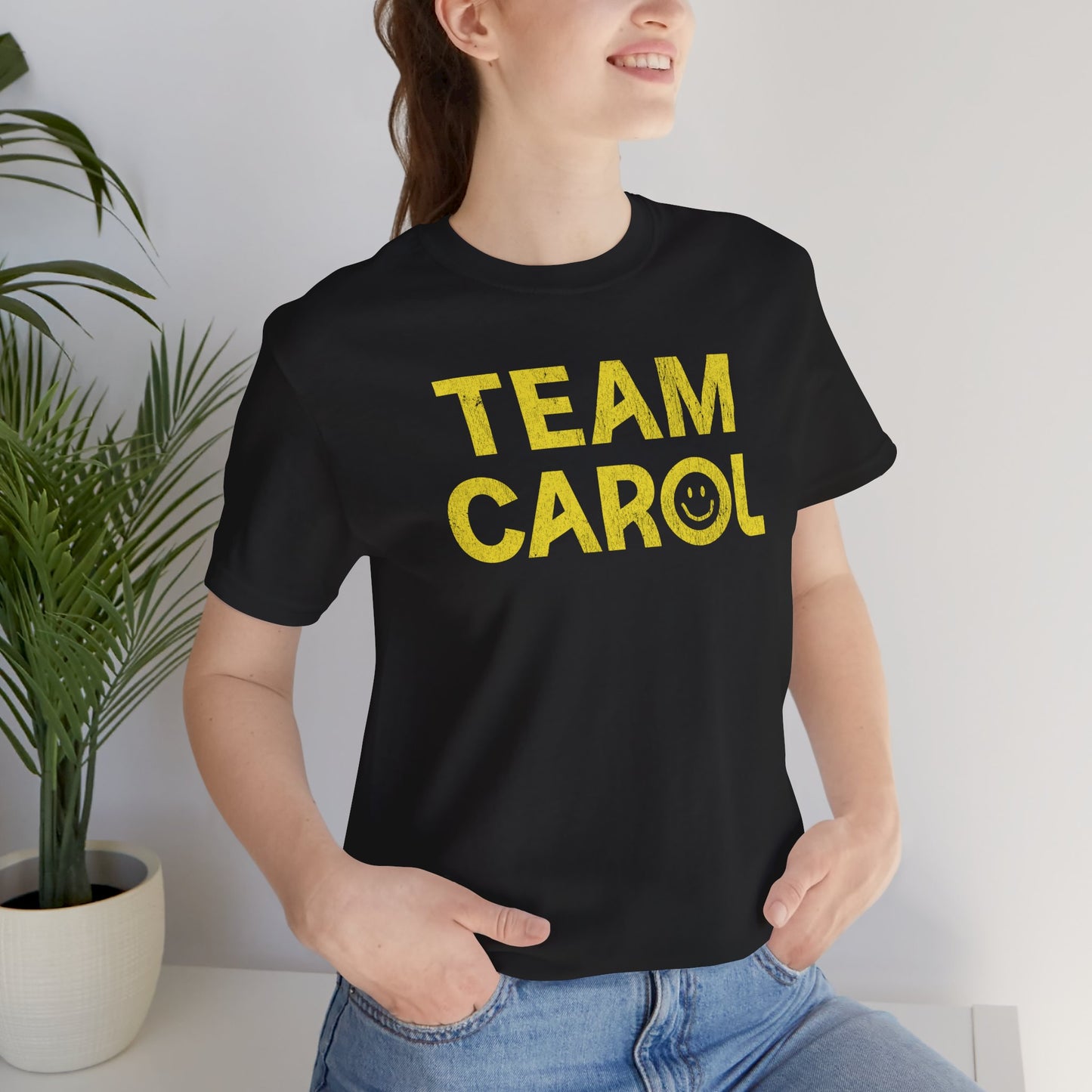 Person wearing Black TEAM CAROL t-shirt inspired by Pluribus with vintage distressed smiley face graphic in yellow, seated near a plant