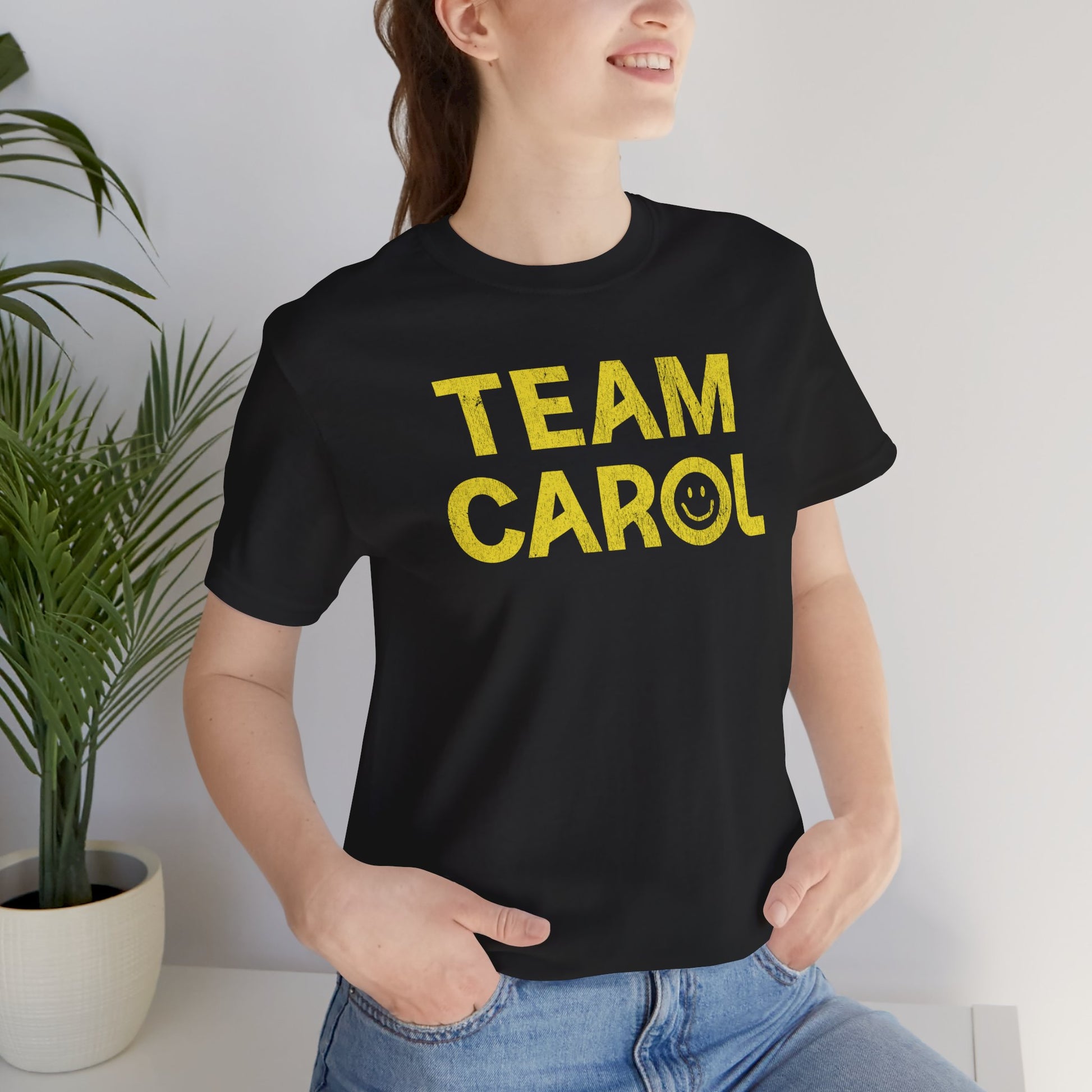 Person wearing Black TEAM CAROL t-shirt inspired by Pluribus with vintage distressed smiley face graphic in yellow, seated near a plant