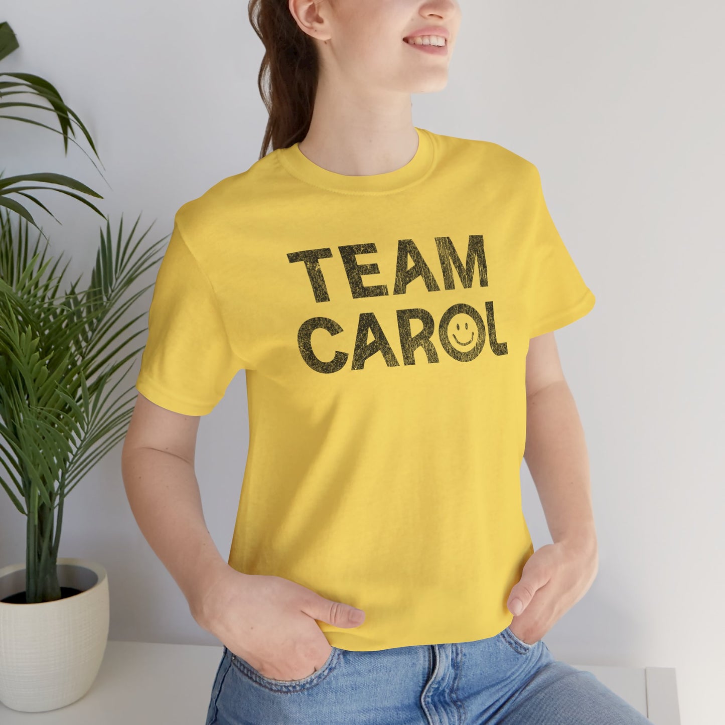 Person wearing yellow TEAM CAROL t-shirt inspired by Pluribus with vintage distressed smiley face graphic in black, seated near a plant