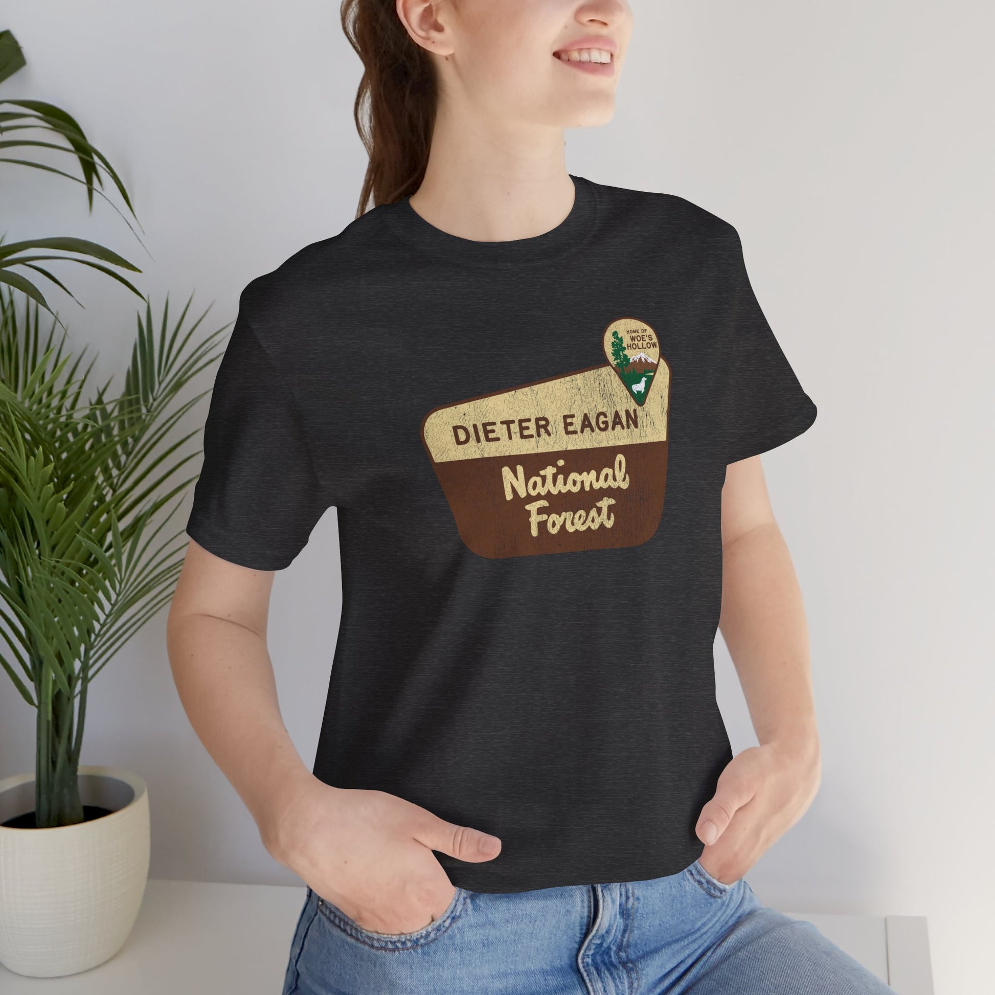 Female model wearing a dark gray heather colored soft Bella+Canvas short-sleeve t-shirt with the logo graphic of the Dieter Eagan National Forest inspired by the TV show Severance in a vintage distressed ink-stamped worn-in style.