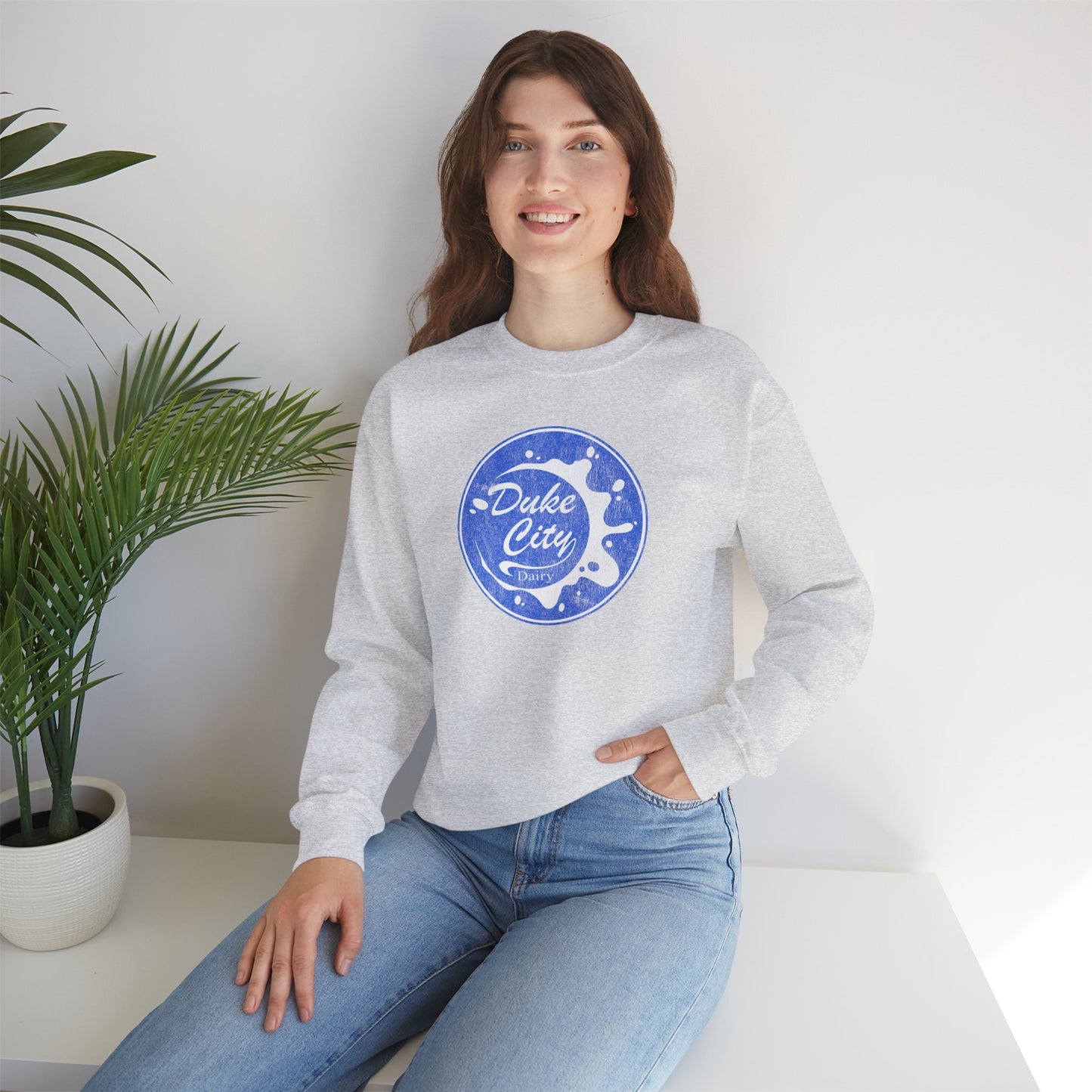 Woman wearing an ash sweatshirt with 'Duke City' logo design from Pluribus, sitting on a white surface with a plant in the background.