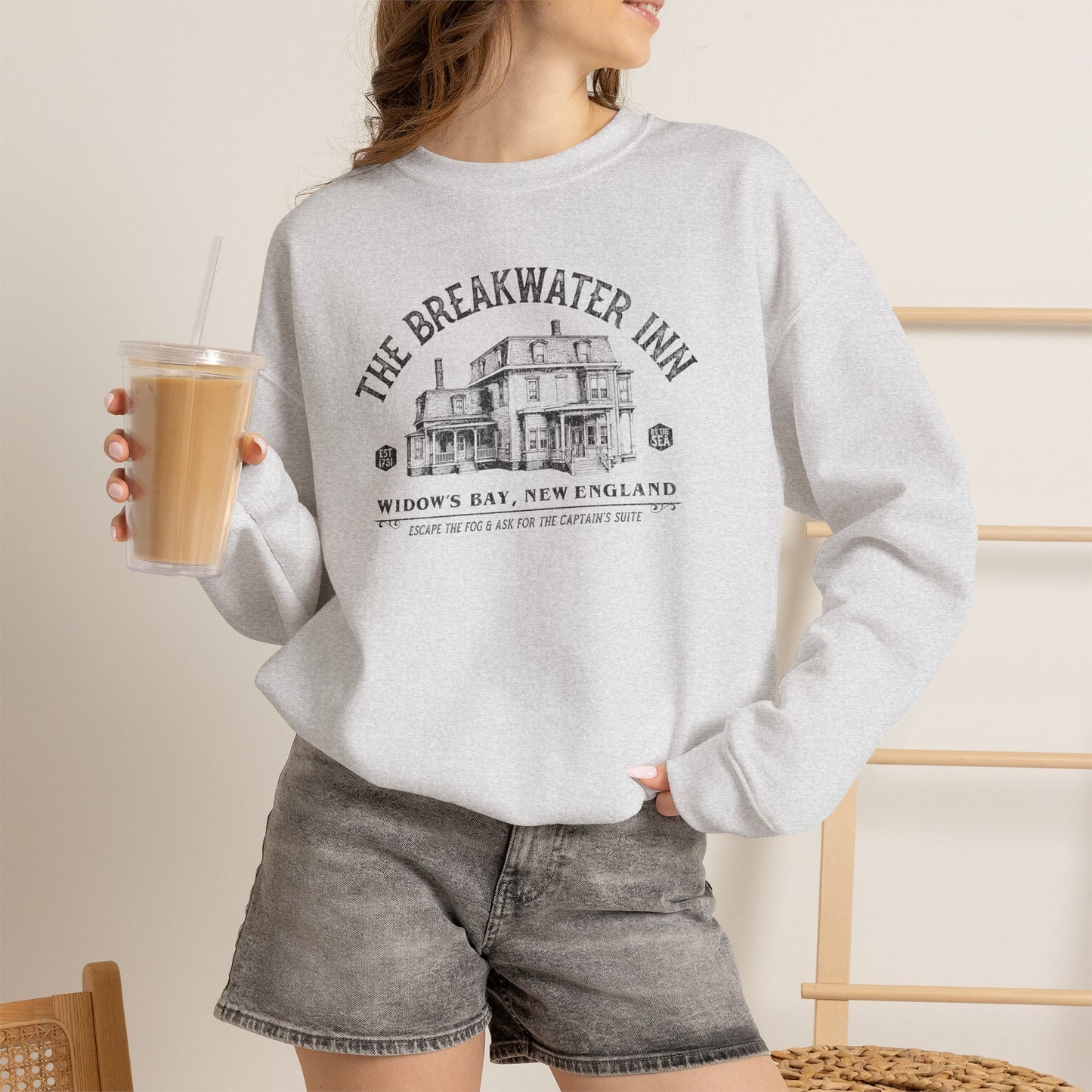 Woman wearing ash crewneck sweatshirt with Breakwater Inn Widow's Bay New England hotel graphic