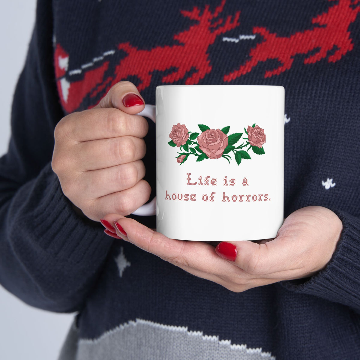 Person holding white mug with pink roses and Life is a house of horrors in cross stitch, inspired by Ruth in Widow's Bay