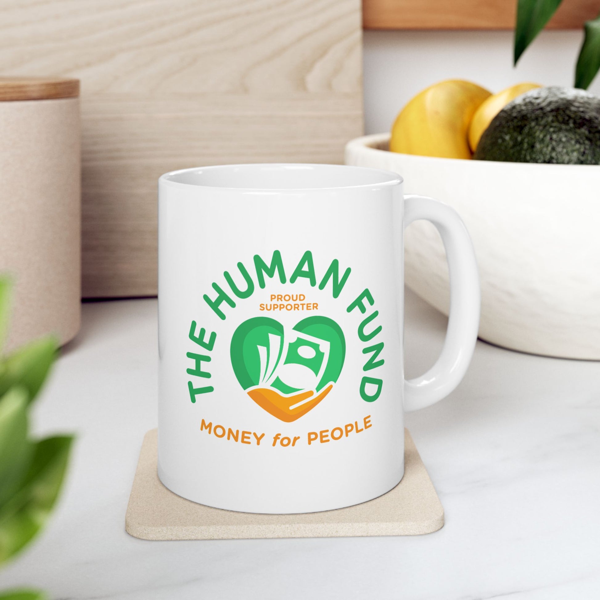 White mug with 'The Human Fund' logo on a white surface with plants and a plant pot in the background.