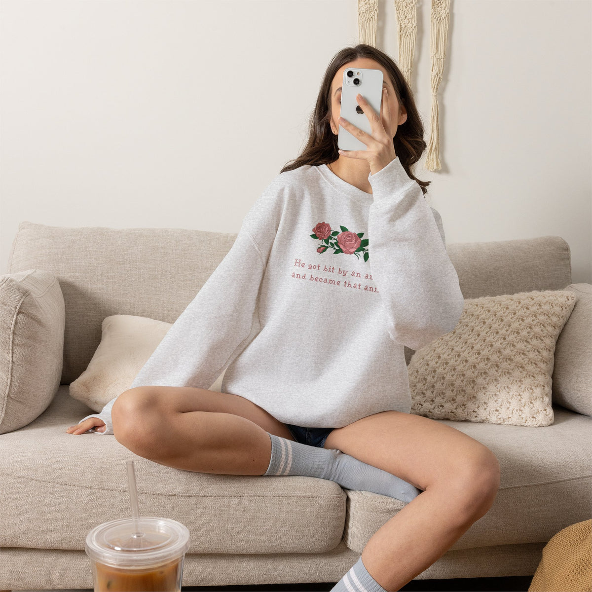 A woman wearing a heather ash crewneck sweatshirt with a graphic of pink roses and the text 'He got bit by an animal, and became that animal.' on a couch setting.