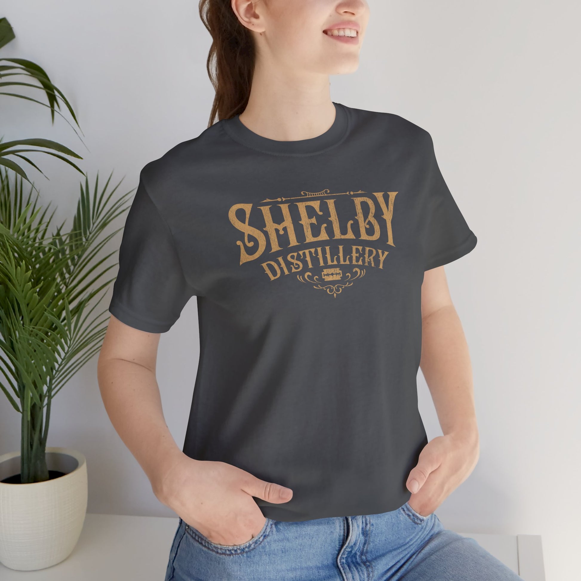 Female model wearing a asphalt gray Soft Bella+Canvas 3001 short-sleeve t-shirt featuring the Shelby Company Limited Distillery Logo Graphic inspired by the TV show and movie Peaky Blinders in a vintage distressed style, sold by ScriptedBrand.