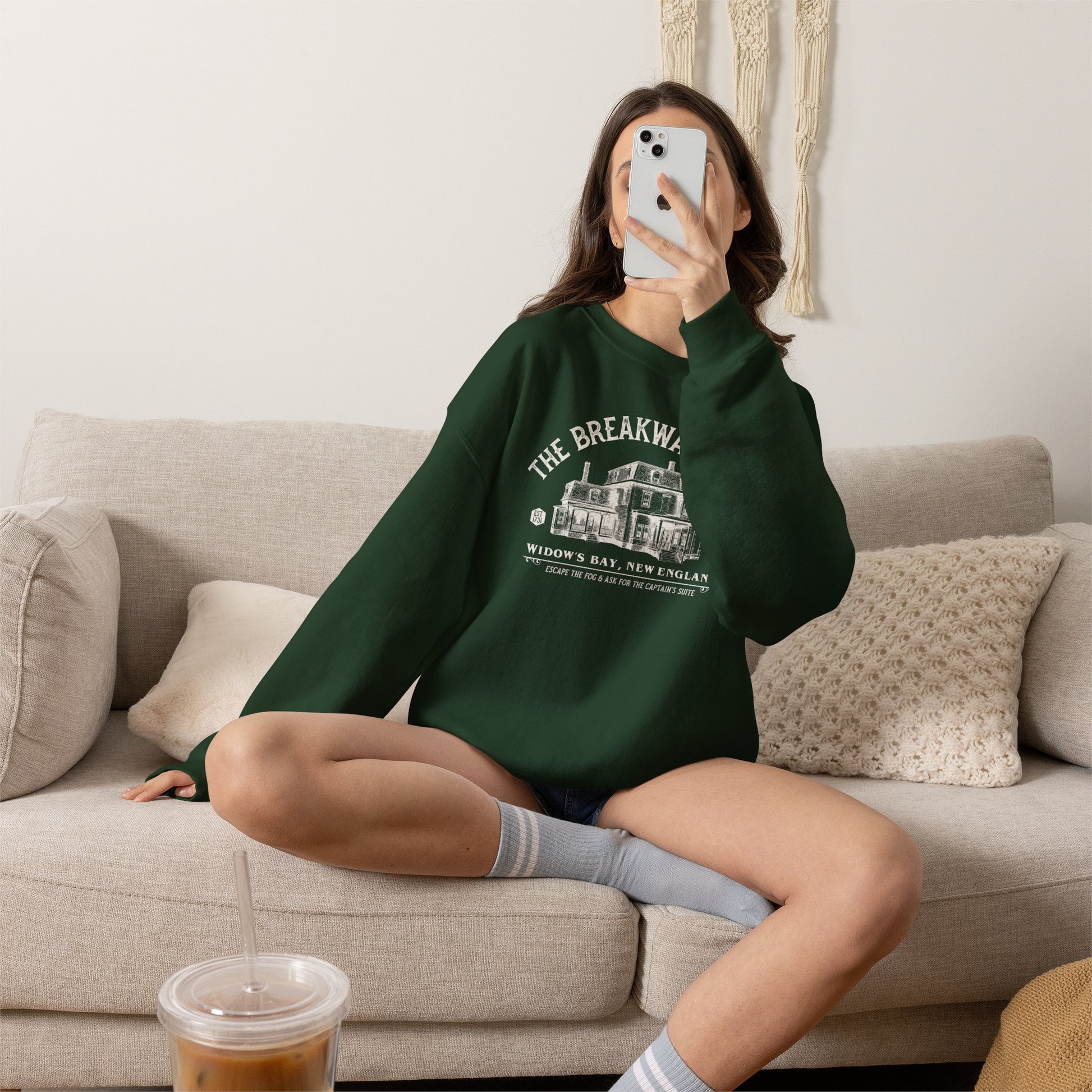Woman on couch wearing forest green crewneck sweatshirt with Breakwater Inn Widow's Bay New England hotel graphic