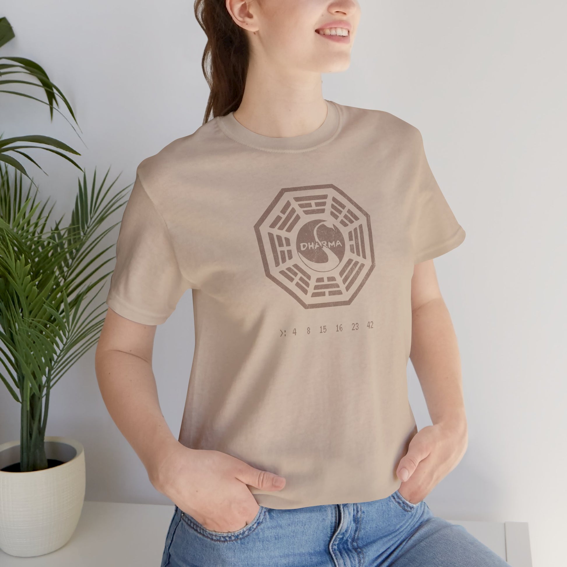 Female model wearing a Tan short-sleeve t-shirt featuring the Dharma Initiative swan station logo graphic from the TV show LOST in a vintage distressed style.