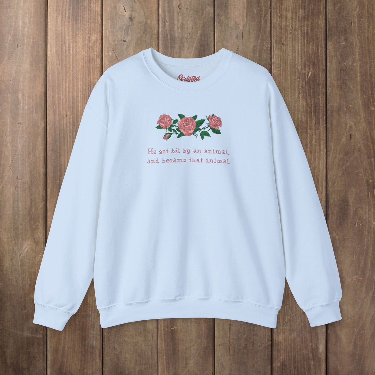 A light blue crewneck sweatshirt with a graphic of pink roses and green leaves on the chest, accompanied by the text 'He hit by an animal, and became that animal' in pink. The sweatshirt is laid flat on a rustic wooden plank background.