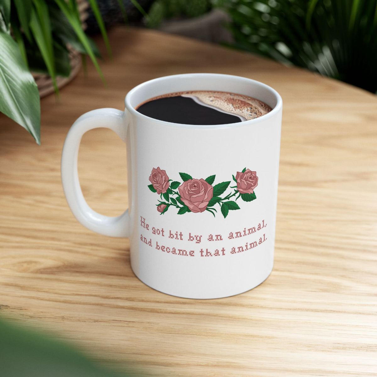 White ceramic mug with a printed design of pink roses and green leaves, featuring the text 'He got bit by an animal, and became that animal' in cross stitch style, inspired by Ruth on Widows Bay, on a wood table