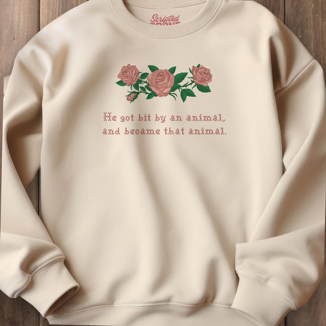 A cream-colored crewneck sweatshirt featuring a graphic of three pink roses with green leaves and the text 'He got bit by an animal, and became that animal.' in a reddish-brown font.