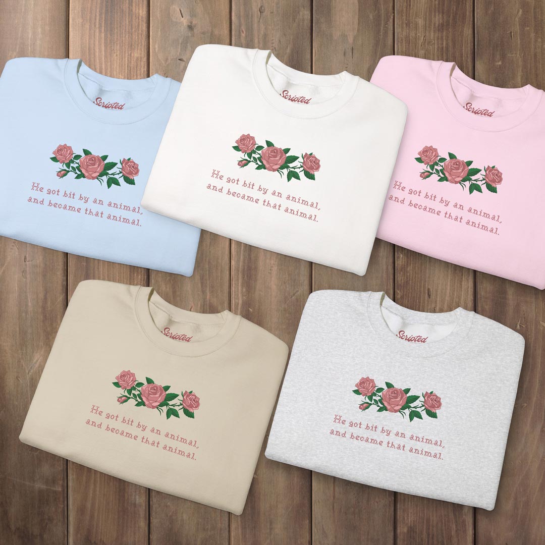 Five folded short-sleeve t-shirts in light blue, cream, pink, beige, and heather gray, each printed with a pink rose design and the phrase 'He got hit by an animal, and became that animal.' on a dark wood background.