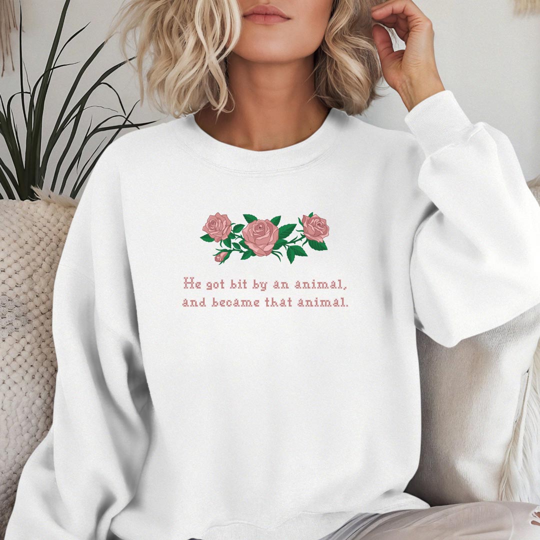 A woman wearing a white crewneck sweatshirt with a graphic of pink roses and the text 'He got bit by an animal, and became that animal.' on a cozy indoor couch setting.