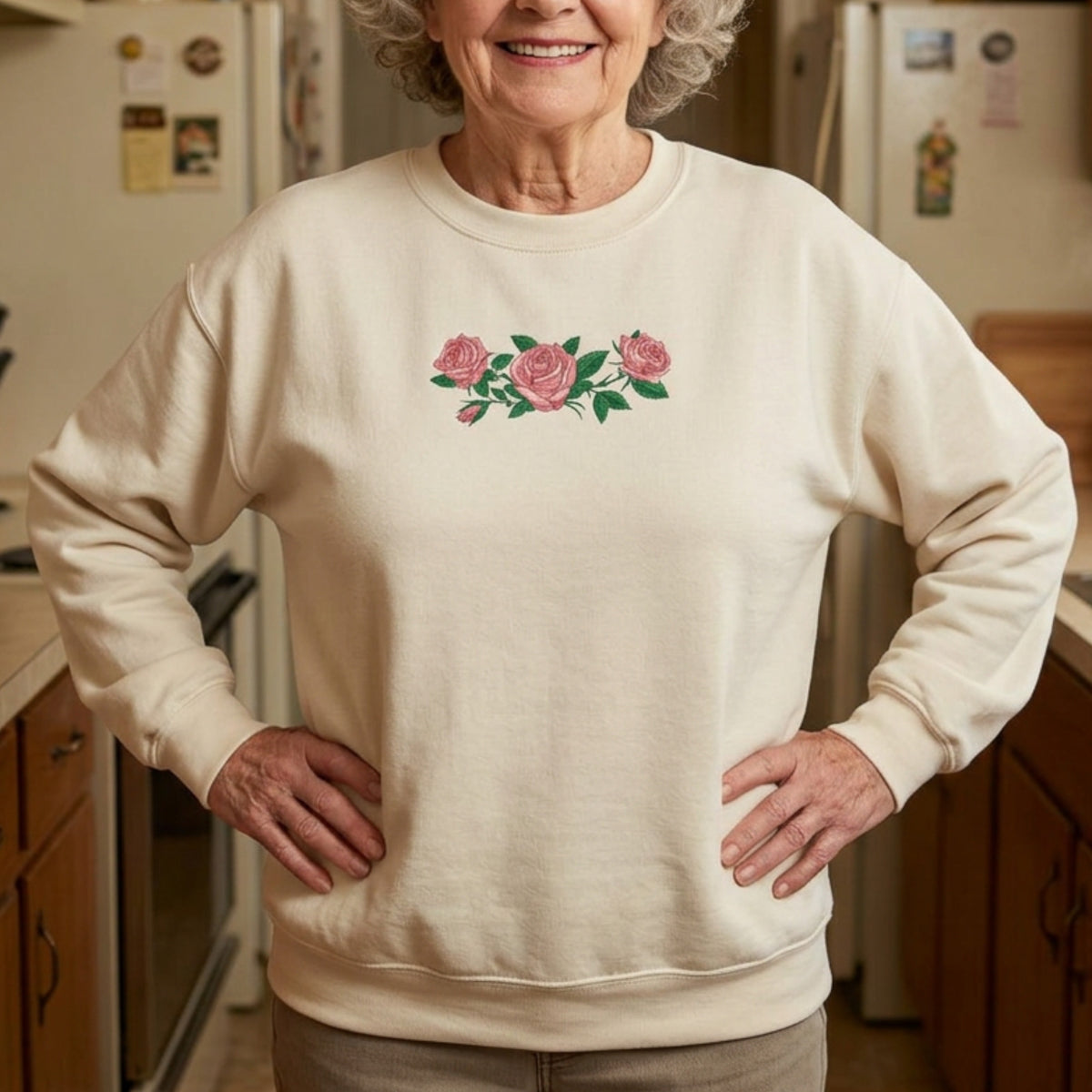An older woman standing in a kitchen wearing a tan crewneck sweatshirt with three pink roses and green leaves printed across the chest, inspired by Ruth of Widow's Bay.