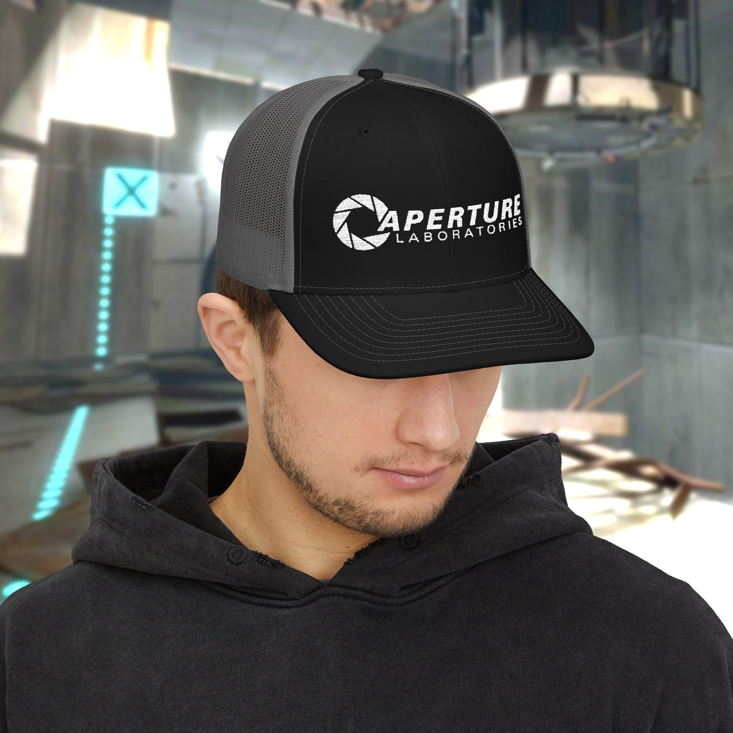 Male model wearing a black/charcoal colored Richardson 112 Snapback Trucker Cap featuring the embroidered logo of the fictional company Aperture Laboratories from the video game Portal and Portal 2, sold by ScriptedBrand. 