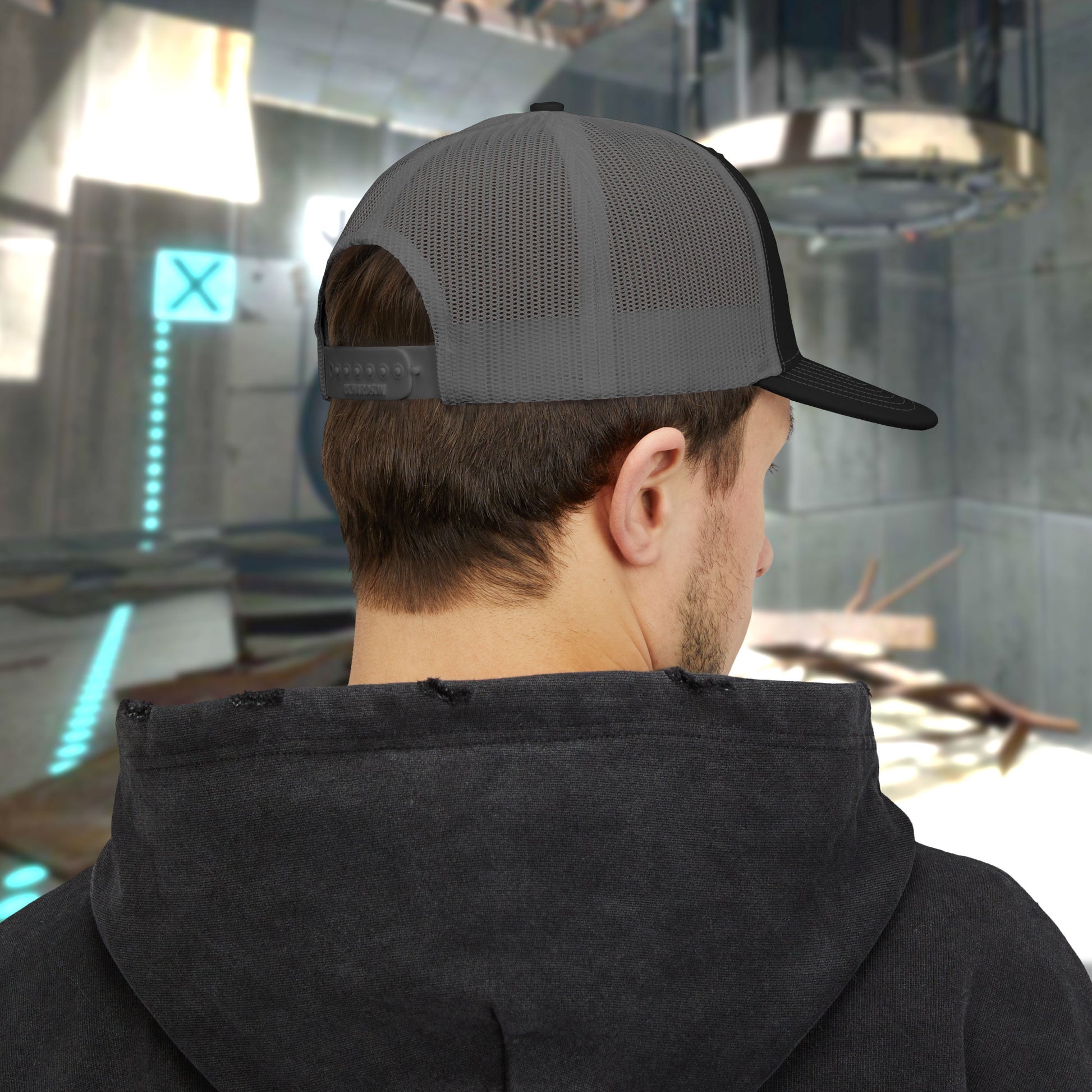 Back view of a male model wearing a black/charcoal colored Richardson 112 Snapback Trucker Cap featuring the embroidered logo of the fictional company Aperture Laboratories from the video game Portal and Portal 2, sold by ScriptedBrand. 