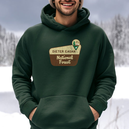 Male model wearing a Forest green colored Soft Unisex Hoodie Heavy Blend Hooded Sweatshirt featuring the logo graphic of the Dieter Eagan National Forest inspired by the TV show Severance in a vintage distressed ink-stamped worn-in style.
