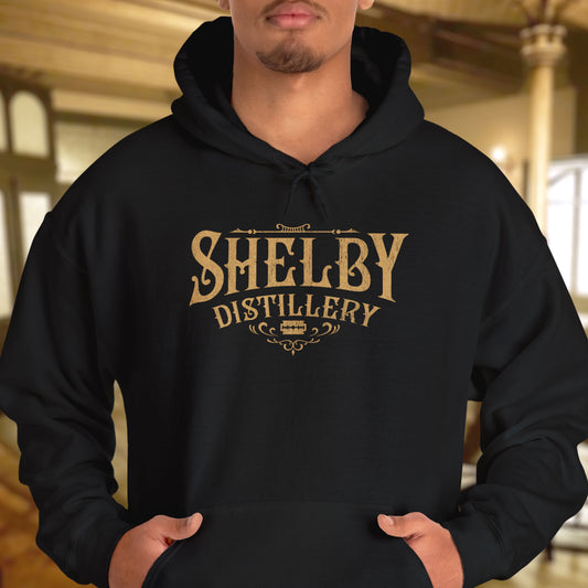 Male model wearing a black Unisex Hoodie Heavy Blend Hooded Sweatshirt featuring the Shelby Company Limited Distillery Logo Graphic inspired by the TV show and movie Peaky Blinders in a vintage distressed ink-stamped worn-in style.