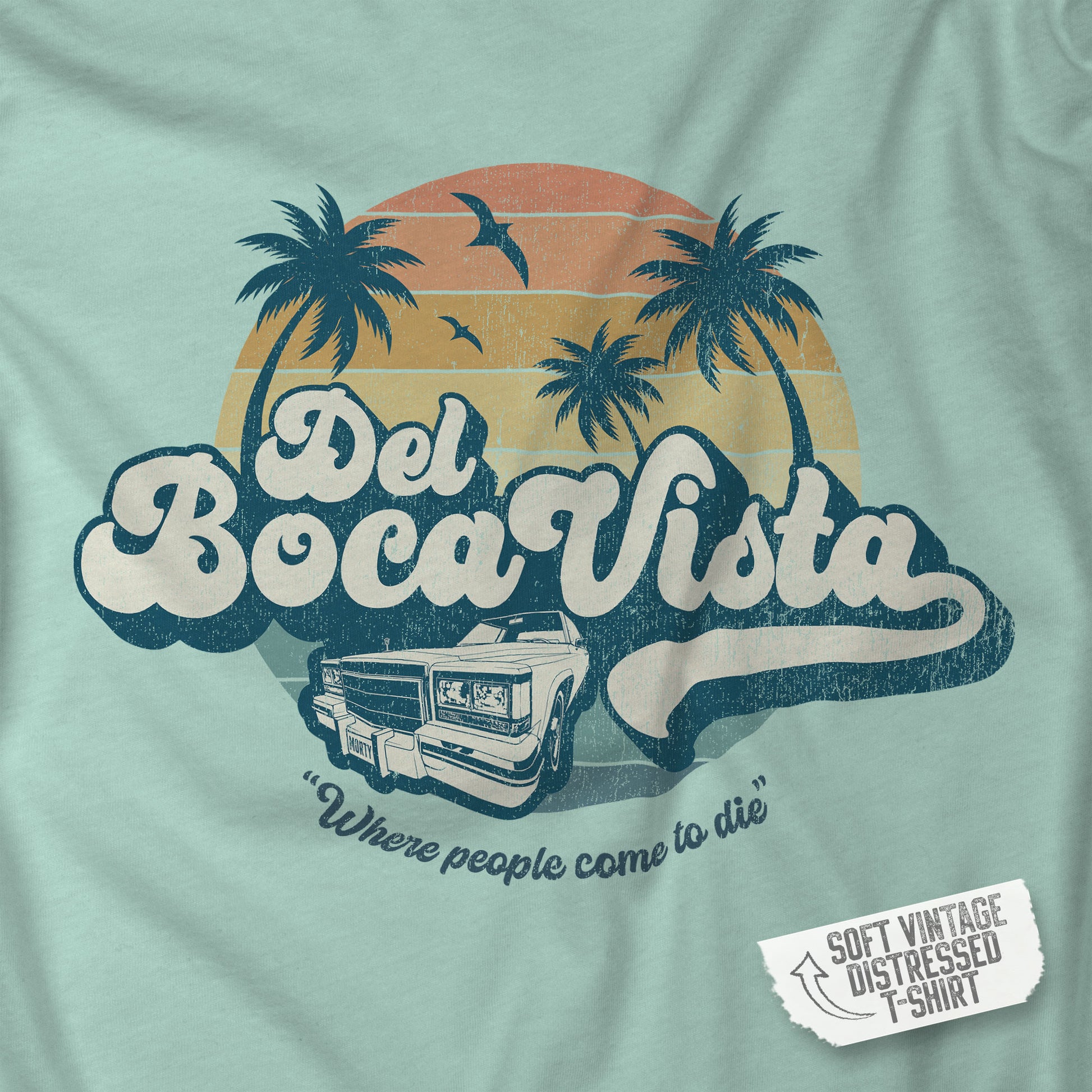 Closeup of a dusty blue Soft Bella+Canvas 3001 short-sleeve t-shirt featuring a Del Boca Vista logo graphic (uniquely designed by ScriptedBrand), including the phrase Where People Come To Die, inspired by the TV show Seinfeld, in a vintage distressed style.