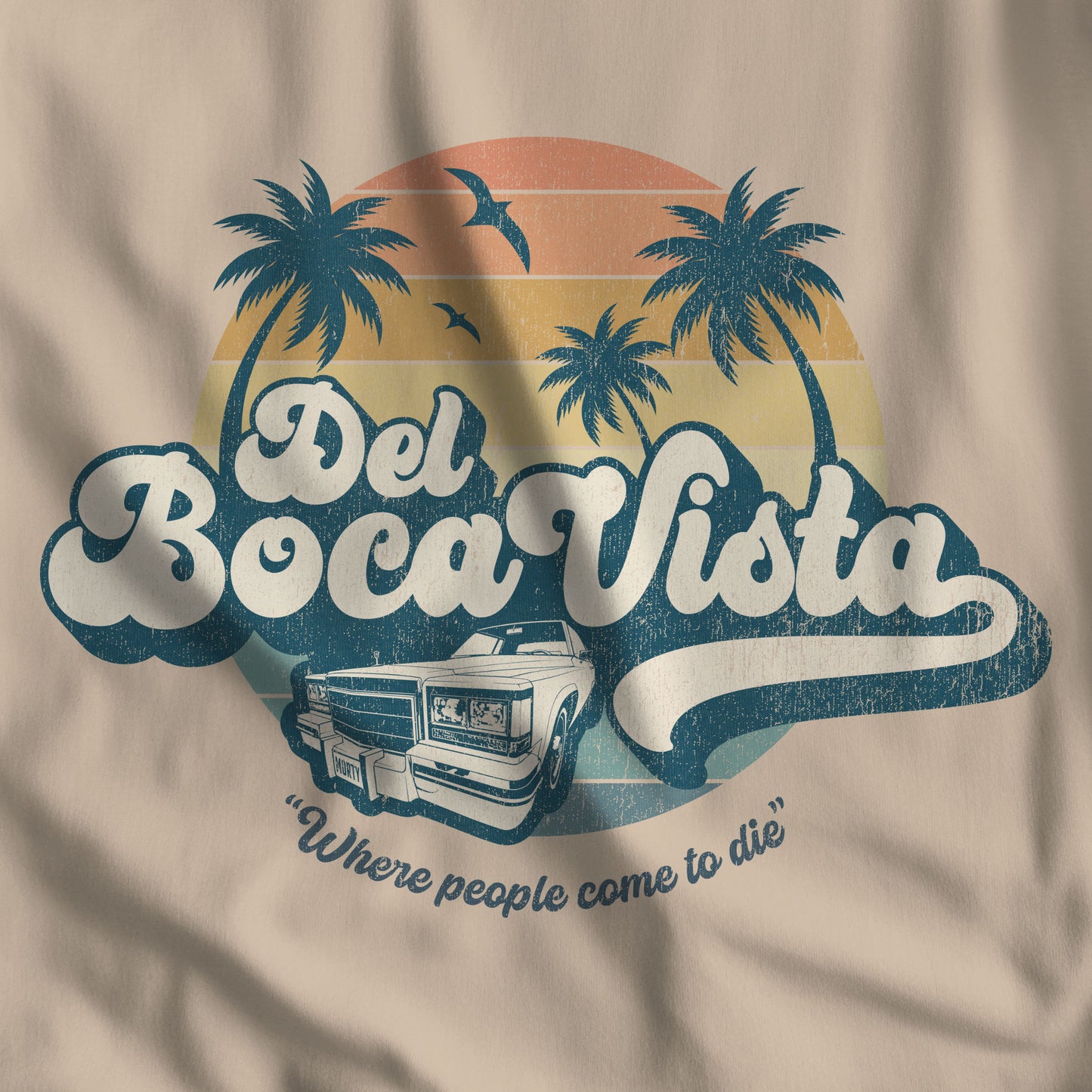 Closeup of tan t-shirt featuring a Del Boca Vista graphic, with phrase Where People Come To Die, inspired by the TV show Seinfeld, in a vintage distressed style.