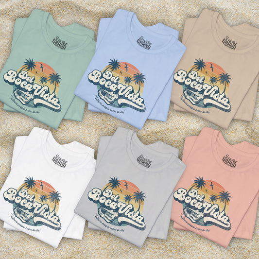 Group of 6 folded Soft Bella+Canvas 3001 short-sleeve t-shirts featuring a Del Boca Vista logo graphic (uniquely designed by ScriptedBrand), including the phrase Where People Come To Die, inspired by the TV show Seinfeld, in a vintage distressed style, laid out on beach sand.