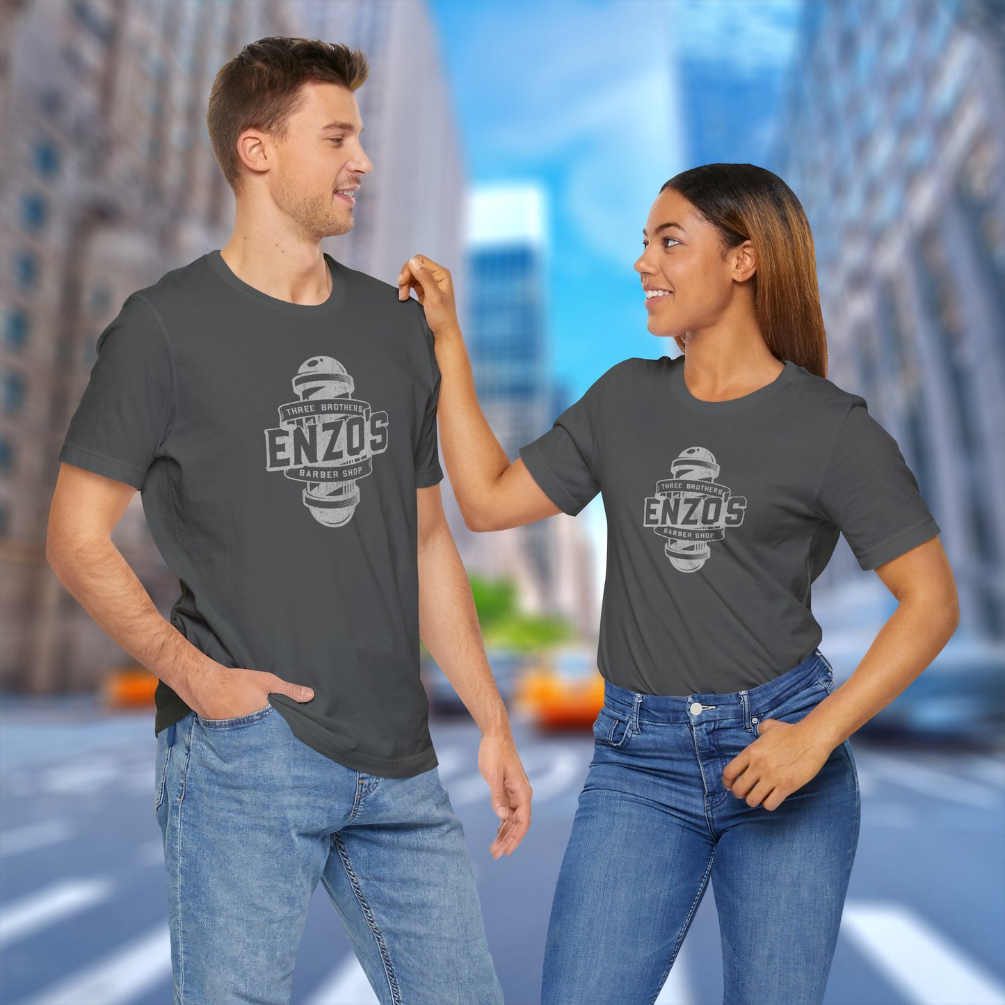 Two people wearing asphalt gray t-shirts with the Enzo's Barber Shop logo graphic inspired by the TV show Seinfeld in a vintage distressed style.