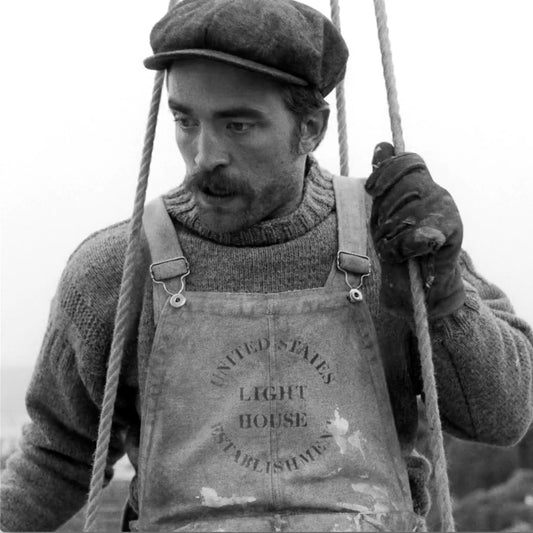 Robert Pattinson wearing overalls with the United States Light House Establishment logo in a scene from the movie The Lighthouse.