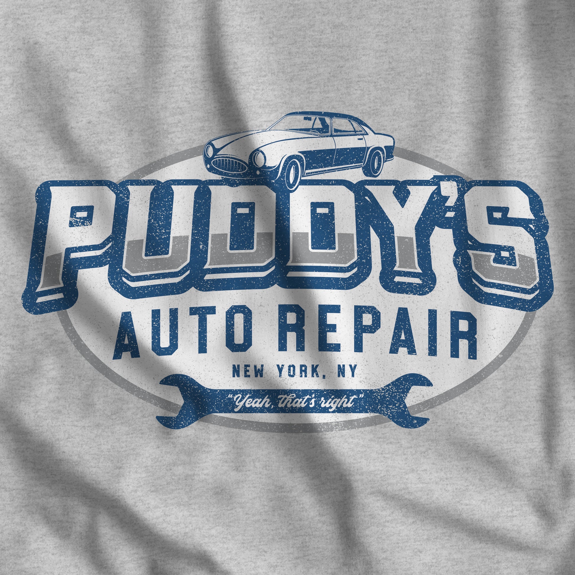 Closeup of heather grey t-shirt with a Puddy's Auto Repair graphic inspired by Seinfeld, in a vintage distressed style.