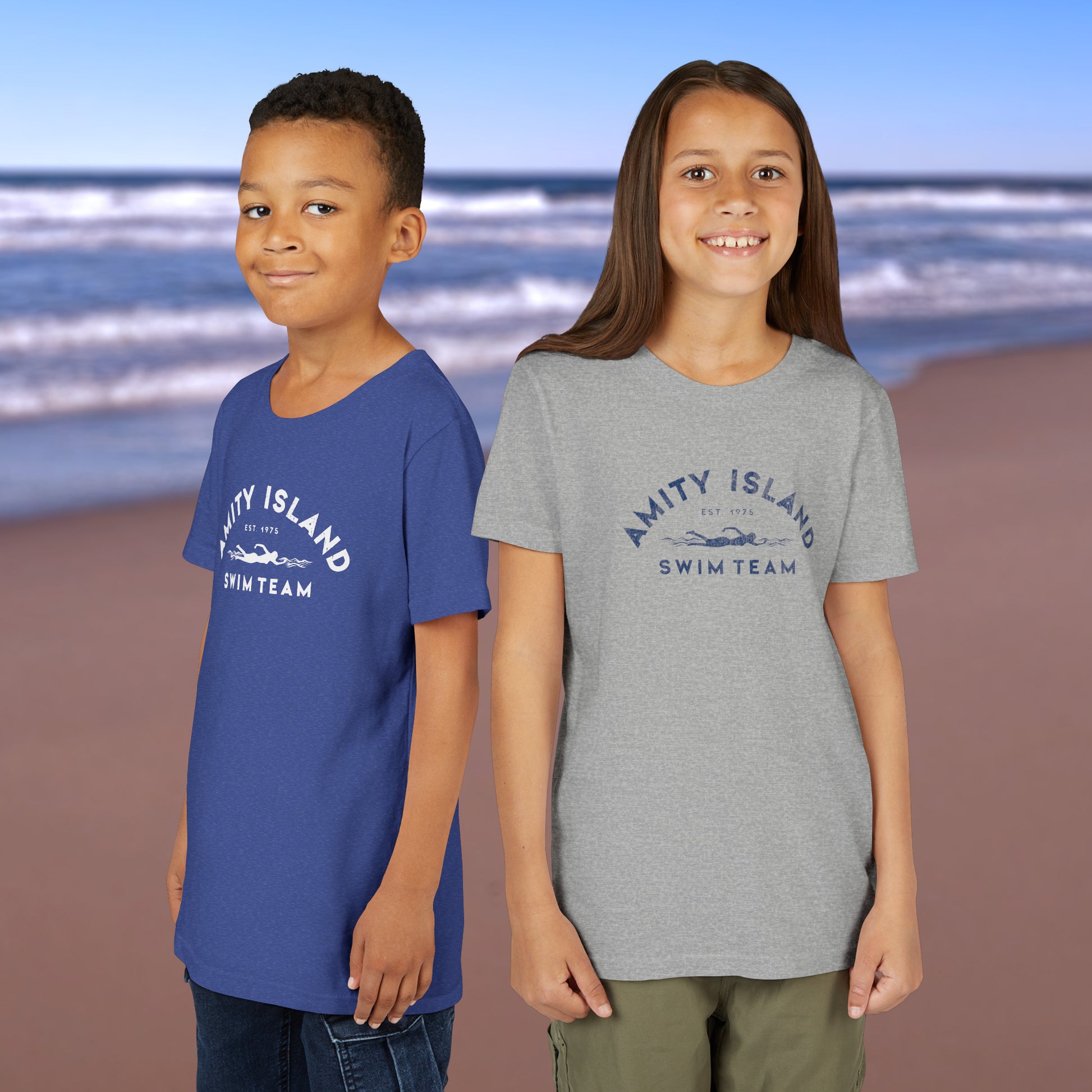2 kid models wearing royal blue and heather grey colored Soft unisex Youth Sizes Bella+Canvas 3001Y short-sleeve t-shirts featuring an Amity Island Swim Team logo graphic, designed by ScriptedBrand, and  inspired by the movie Jaws in a vintage distressed style.