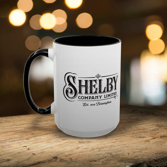 Mockup of a 15 oz White Ceramic Mug with black handle/interior featuring the fictional Shelby Company Limited Logo from the TV show Peaky Blinders printed on both sides. Graphic is designed by ScriptedBrand and includes the text "Est. 1919 Birmingham". Mug is shown placed on a wooden table.