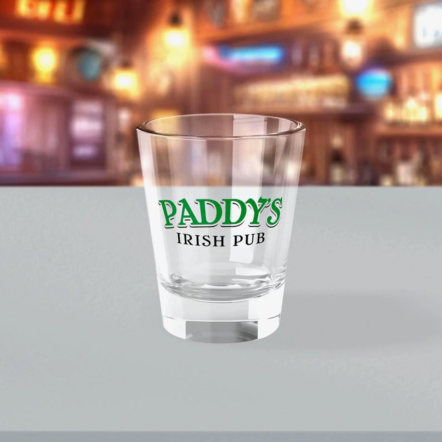 1.5 oz clear Paddy’s Pub shot glass with green logo inspired by It’s Always Sunny in Philadelphia, shown in a bar.