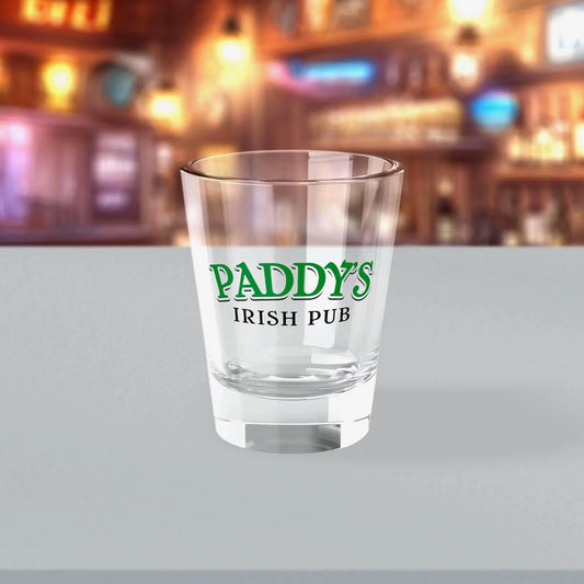 1.5 oz clear Paddy’s Pub shot glass with green logo inspired by It’s Always Sunny in Philadelphia, shown in a bar.