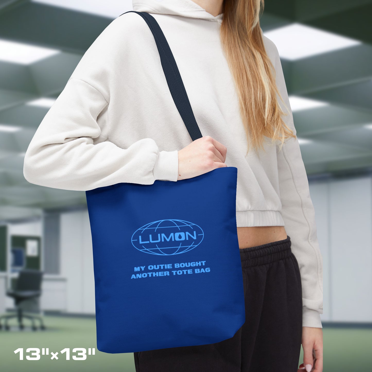 Female model carrying a 13" x 13" Blue Tote Bag featuring the logo of Lumon Industries from the Apple TV Show Severance, along with added customizable text by the customer, which is "MY OUTIE BOUGHT ANOTHER TOTE BAG.".