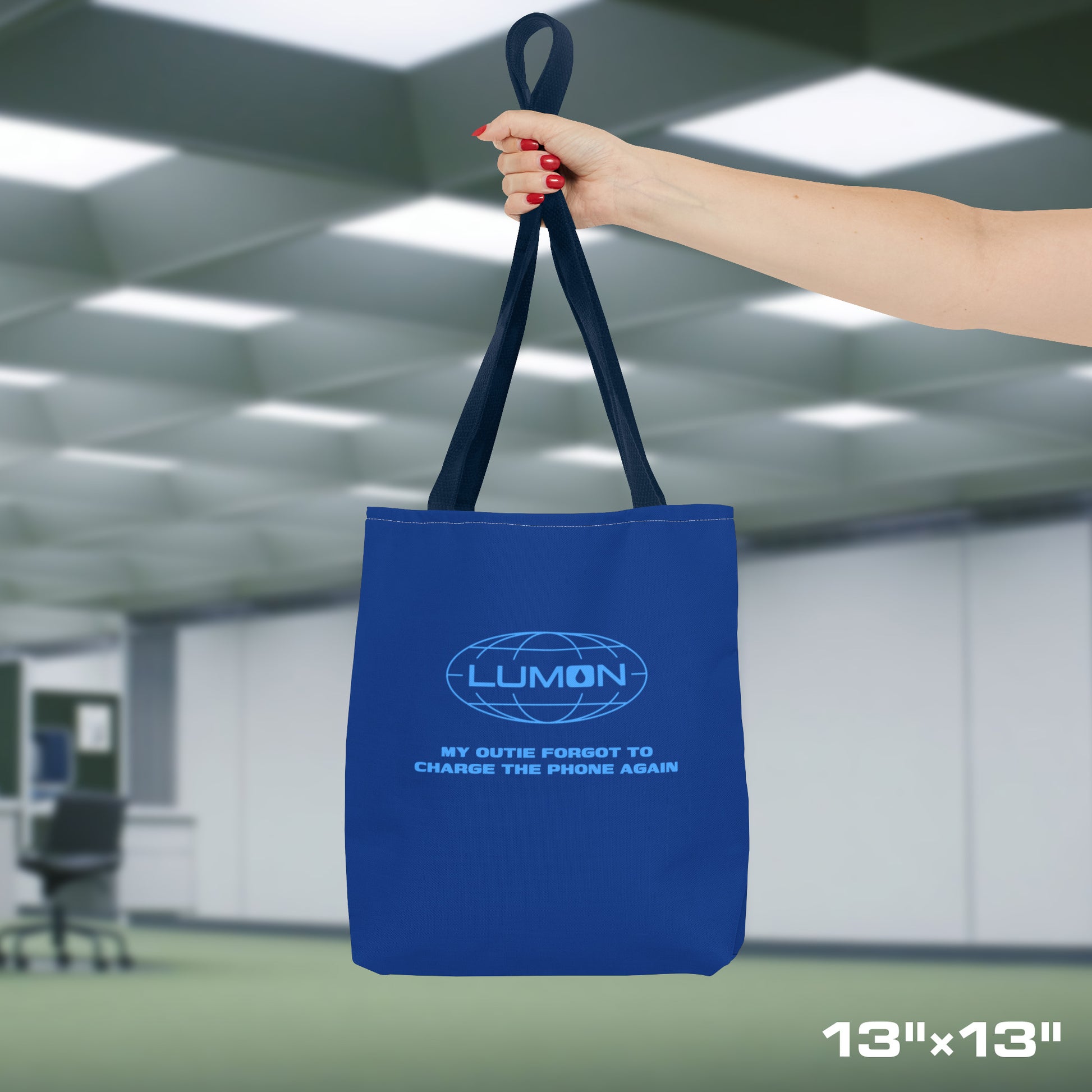 Female model holding a 13" x 13" Blue Tote Bag featuring the logo of Lumon Industries from the Apple TV Show Severance, along with added customizable text by the customer, which is "MY OUTIE FORGOT TO CHARGE THE PHONE AGAIN."