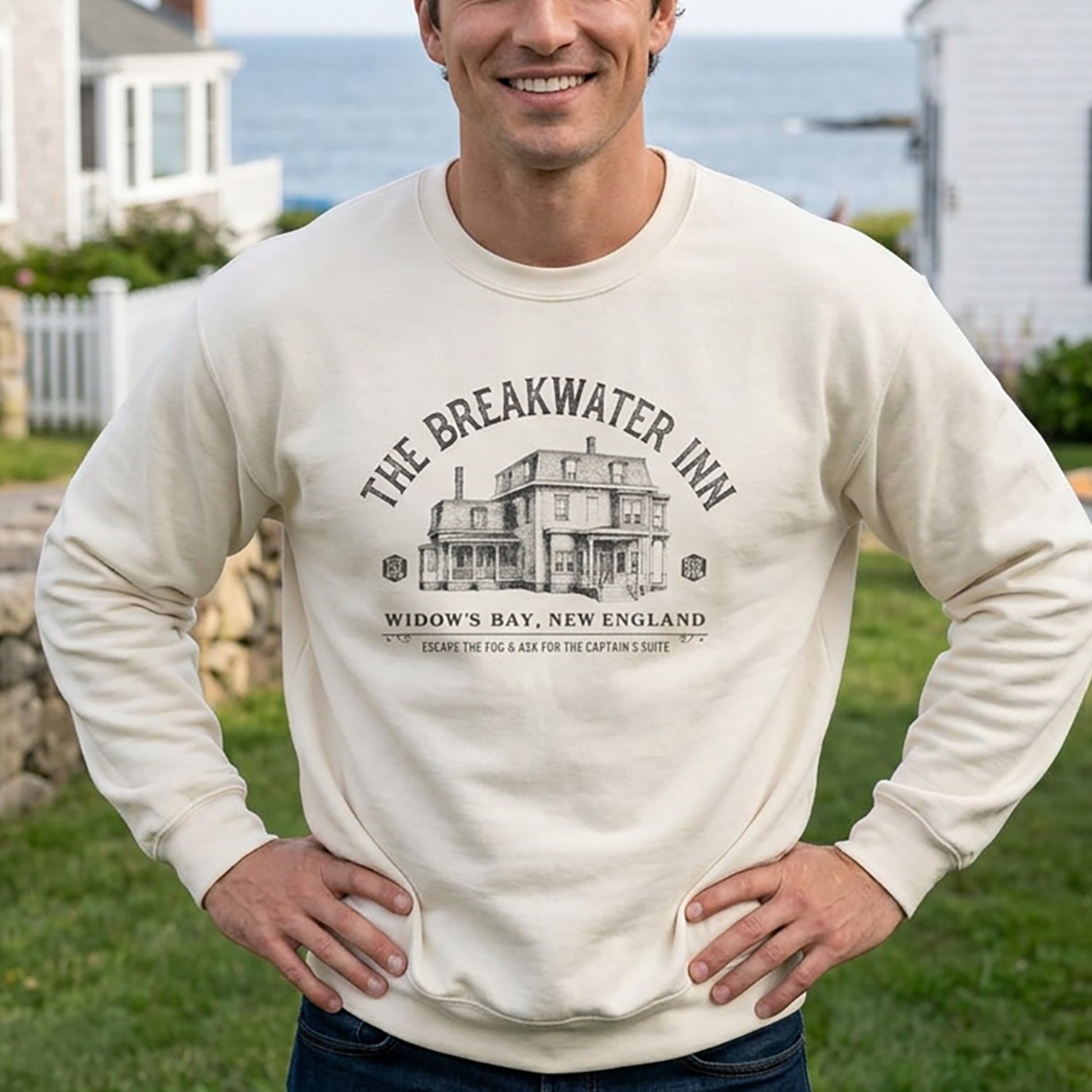 A man wearing a cream-colored crewneck sweatshirt with a black graphic print of 'The Breakwater Inn' in Widow's Bay, New England. The background shows a blurred outdoor coastal scene with grass, houses, and the ocean.